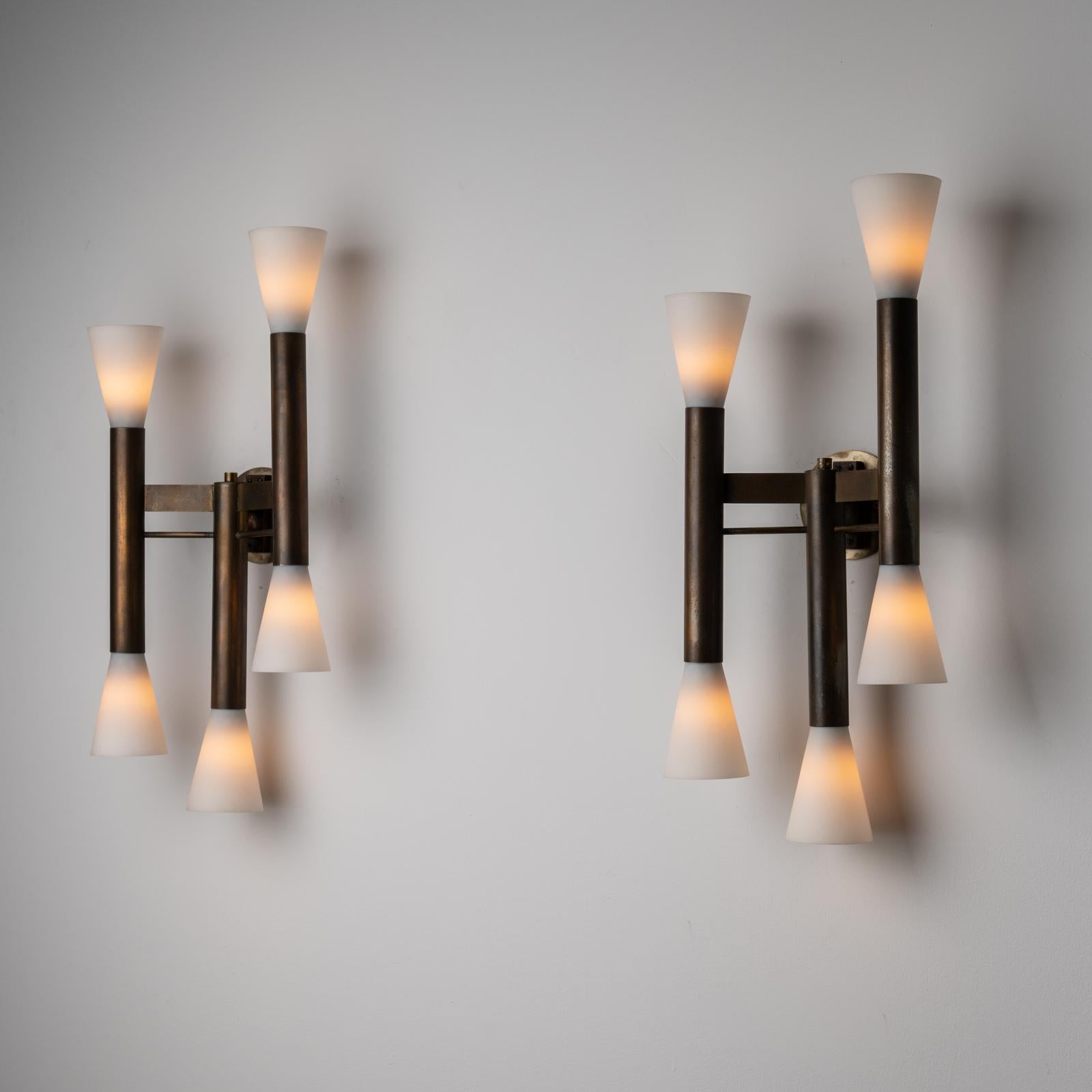 A pair of sconces by Stilnovo. Designed and manufactured in Italy, c. 1960s. Striking torch-style fixtures made up of beautifully aged brass and fluted opal glass shades. Each sconce comprises of three variable tiered armatures. Each stem receives a