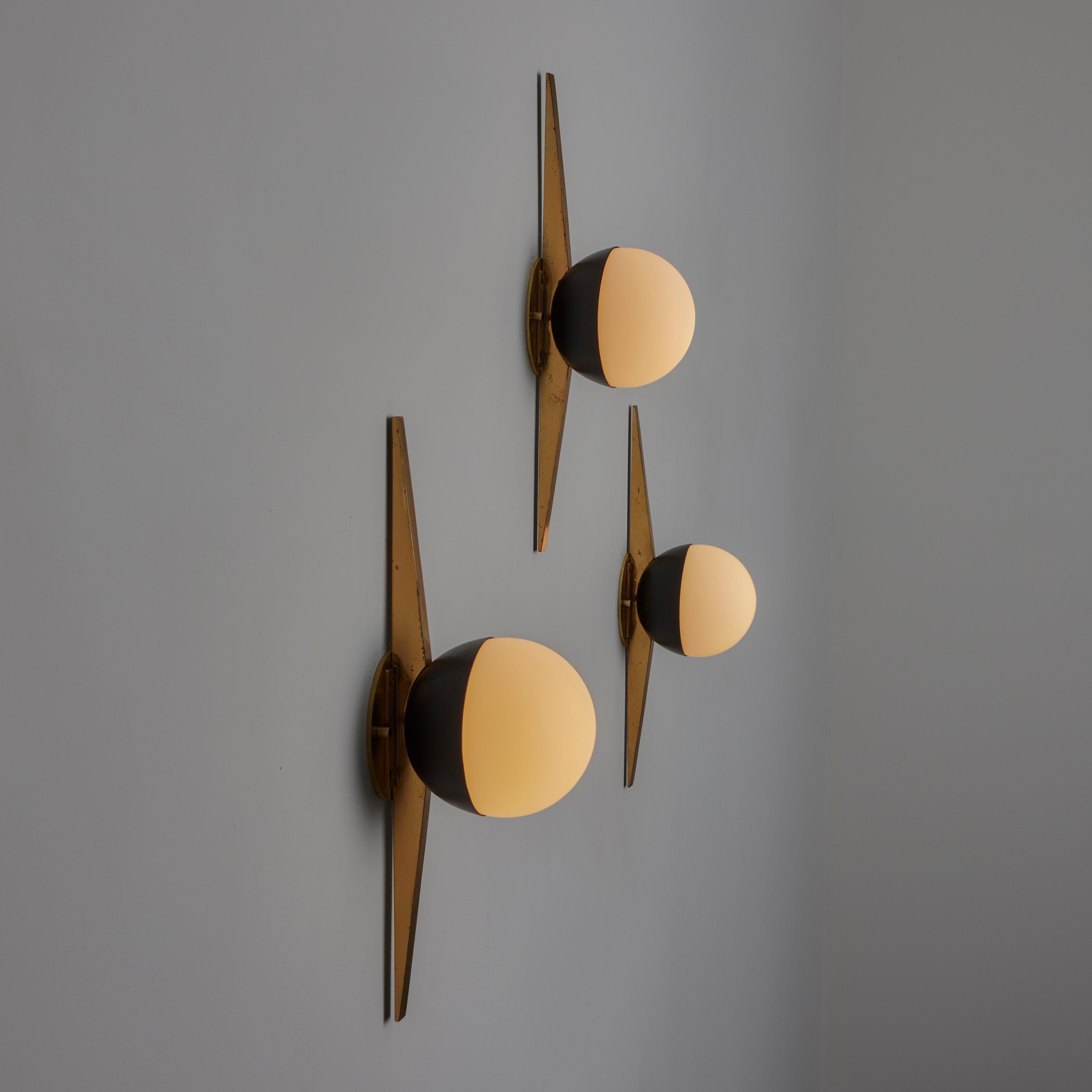Sconces by Stilnovo. Designed and manufactured in Italy, circa the 1950s. Emblem styled wall sconces featuring patinated polished brass extremities equal distant to the center opal glass diffuser. Each sconce holds an e14 socket type, adapted for