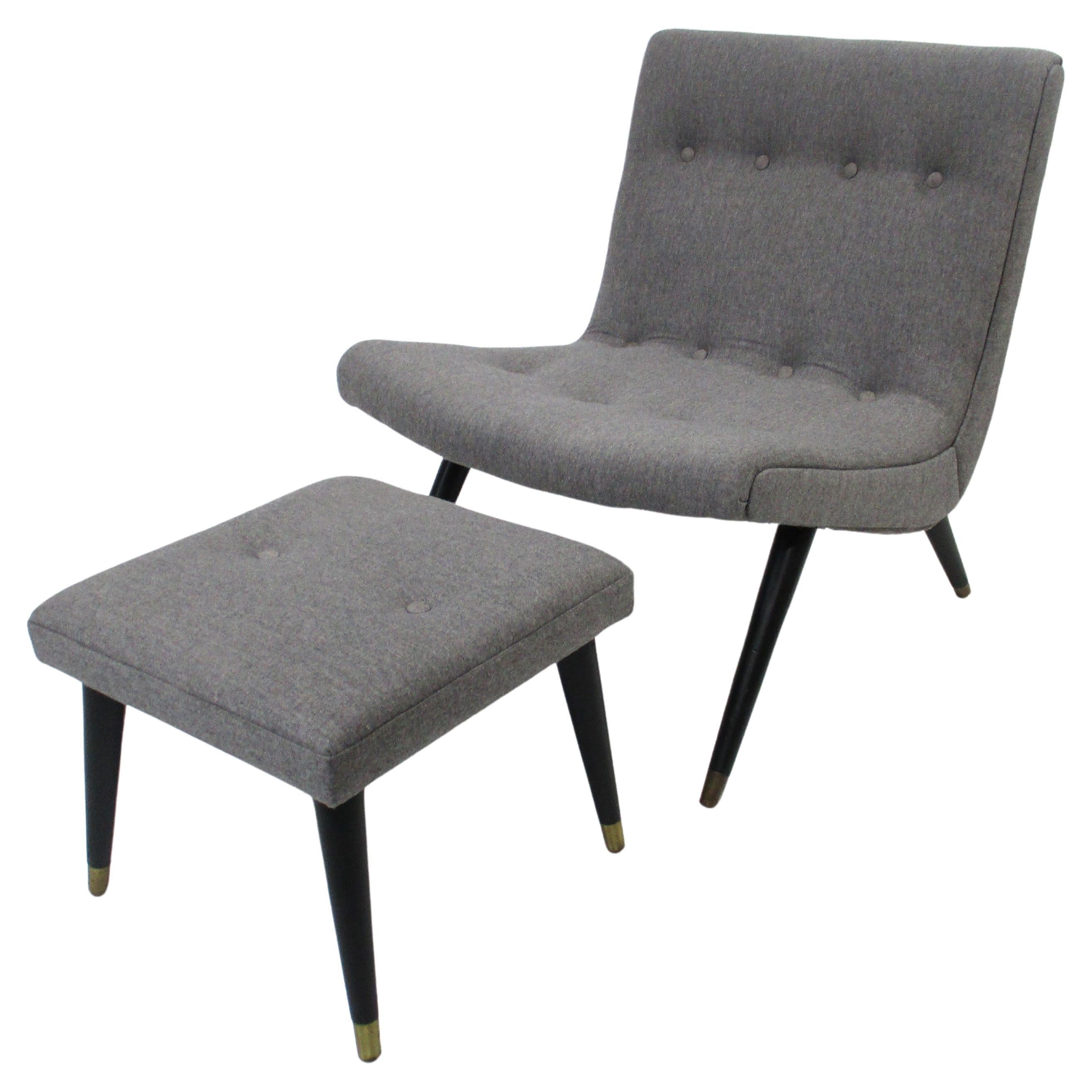 A upholstered scoop styled lounge chair and matching ottoman having  buttons to the top cushions , satin black legs and brass leg tips. Designed in the manner of Milo Baughman and covered in a grey wool flannel styled blend. The ottoman measurement
