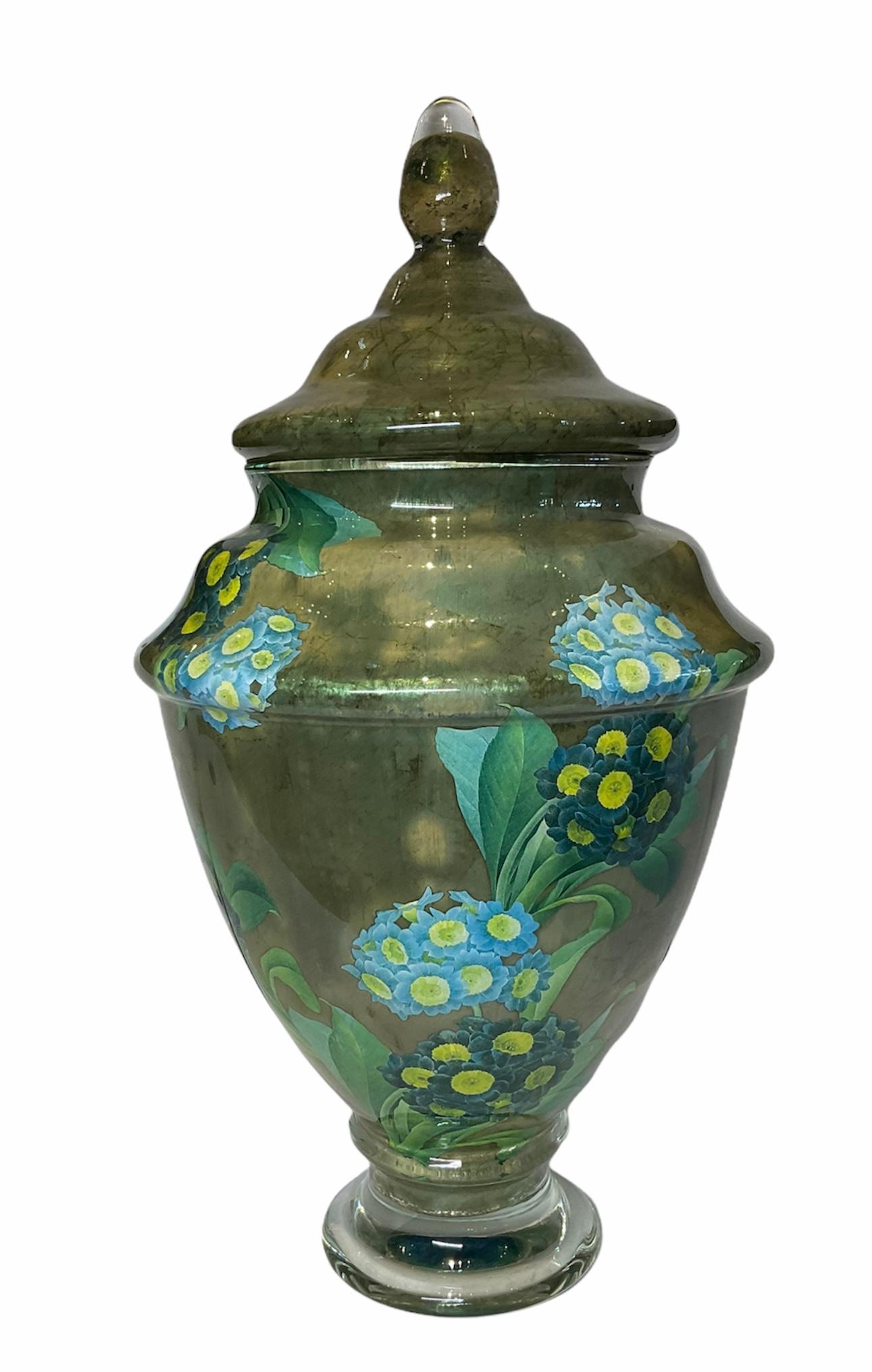 This is a Scott Potter Art Glass lidded urn. It depicts a large urn decoupaged olive green print highlighted with two bouquets of light and dark blue Hydrangeas flowers. This technique was originated in the 18th century and he implemented it with