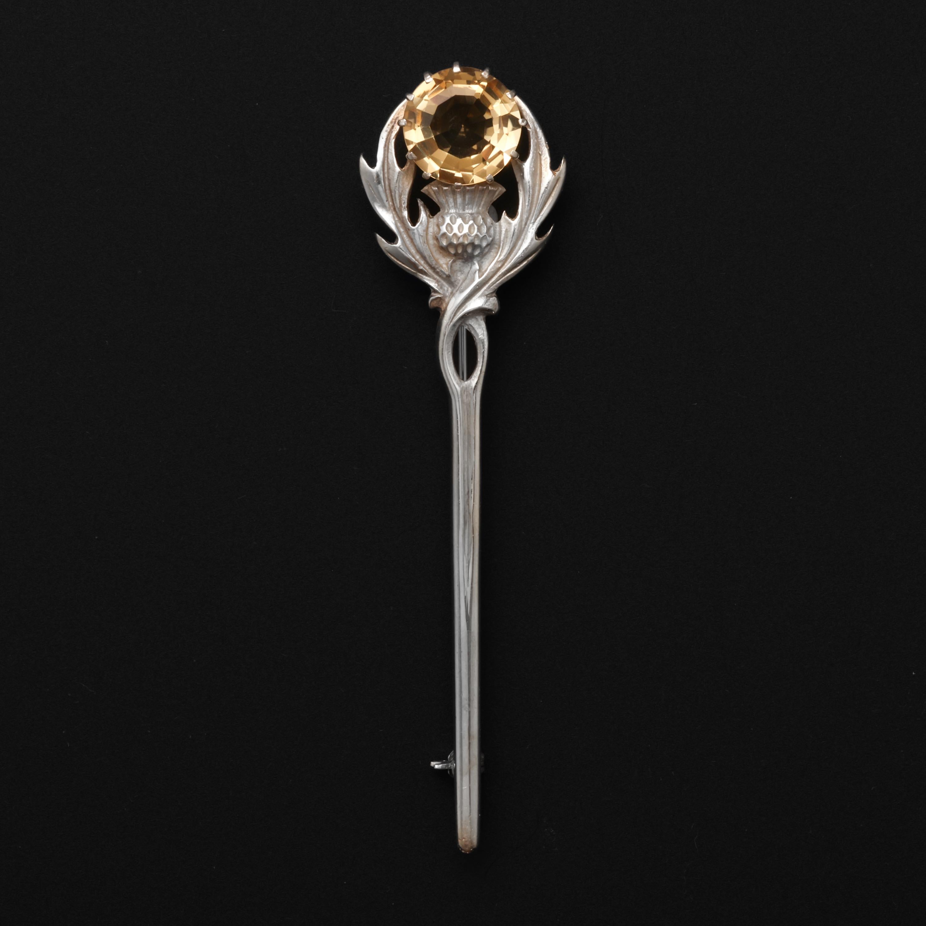 This midcentury (circa 1970) Scottish brooch features a thistle motif with two shaggy leaves holding up a large (12mm) faceted round citrine gemstone. Citrine is found in limited amounts in the Cairngorm Mountains in Scotland. Thus, Cairngorm