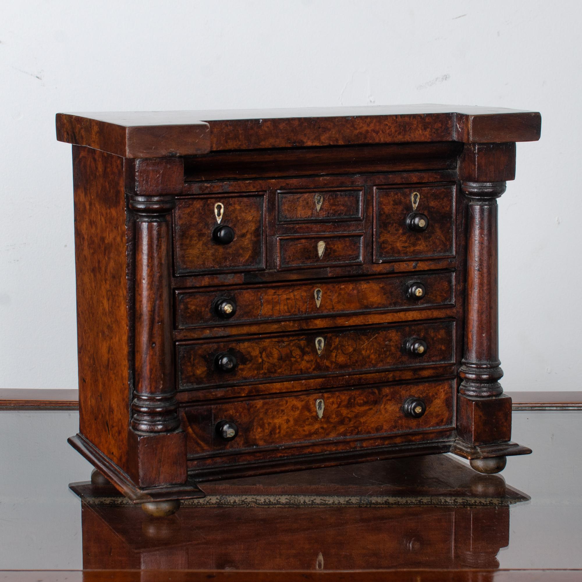 A Scottish miniature chest of drawers in burled wood.  

Drawer inscribed on underside with maker’s name: 

“E.B. McGowan; Bank St, Wigtown; 17th February 1863”

Hidden drawer on top with hole in rear to push open.  

Two small central drawers are