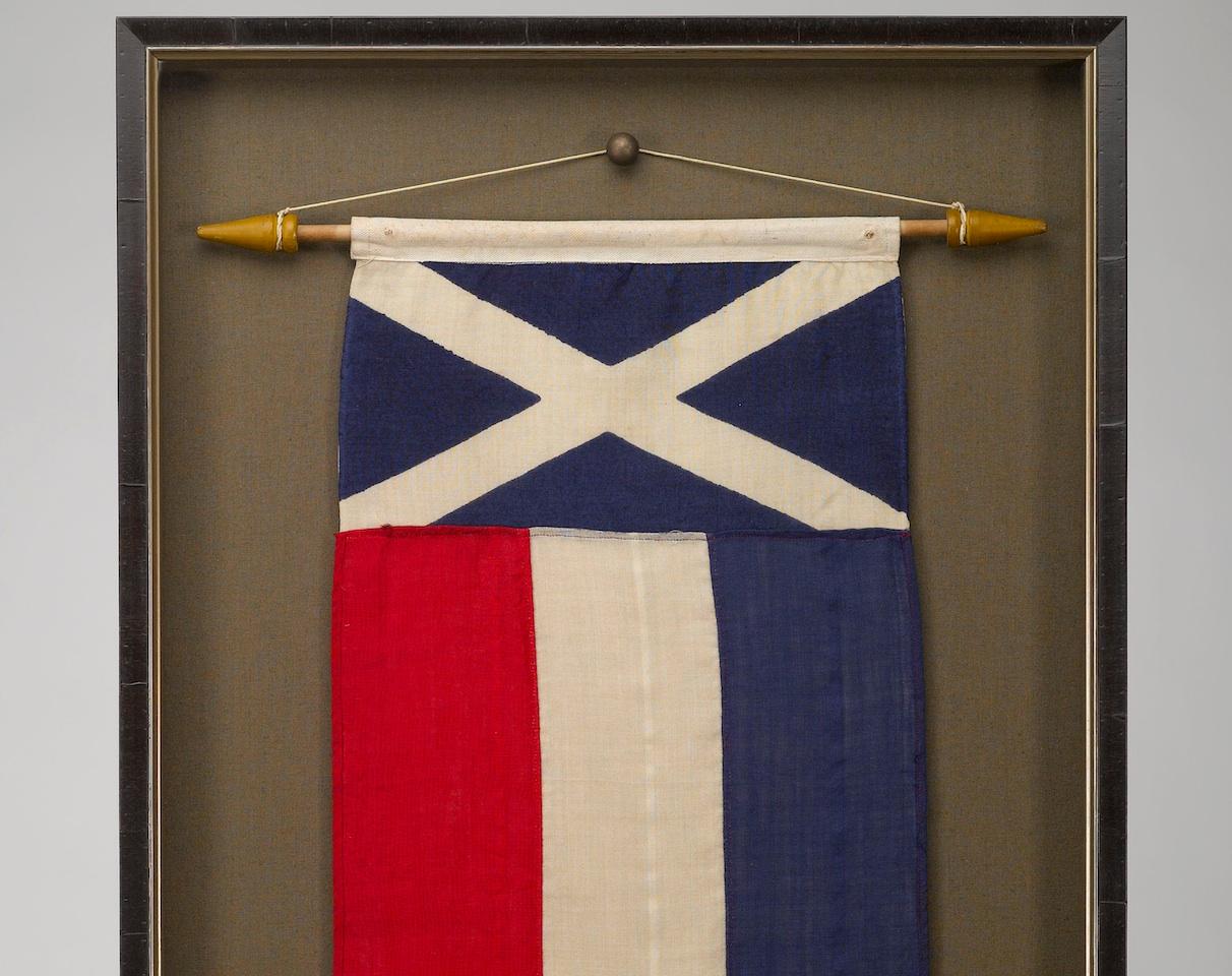 Presented is a Scottish celebratory banner from the coronation of Queen Elizabeth II in 1953. The banner hangs vertically from a wooden dowel. The canton is composed of the x-shaped White Cross of St. Andrew on a blue field. Called the Saltire or