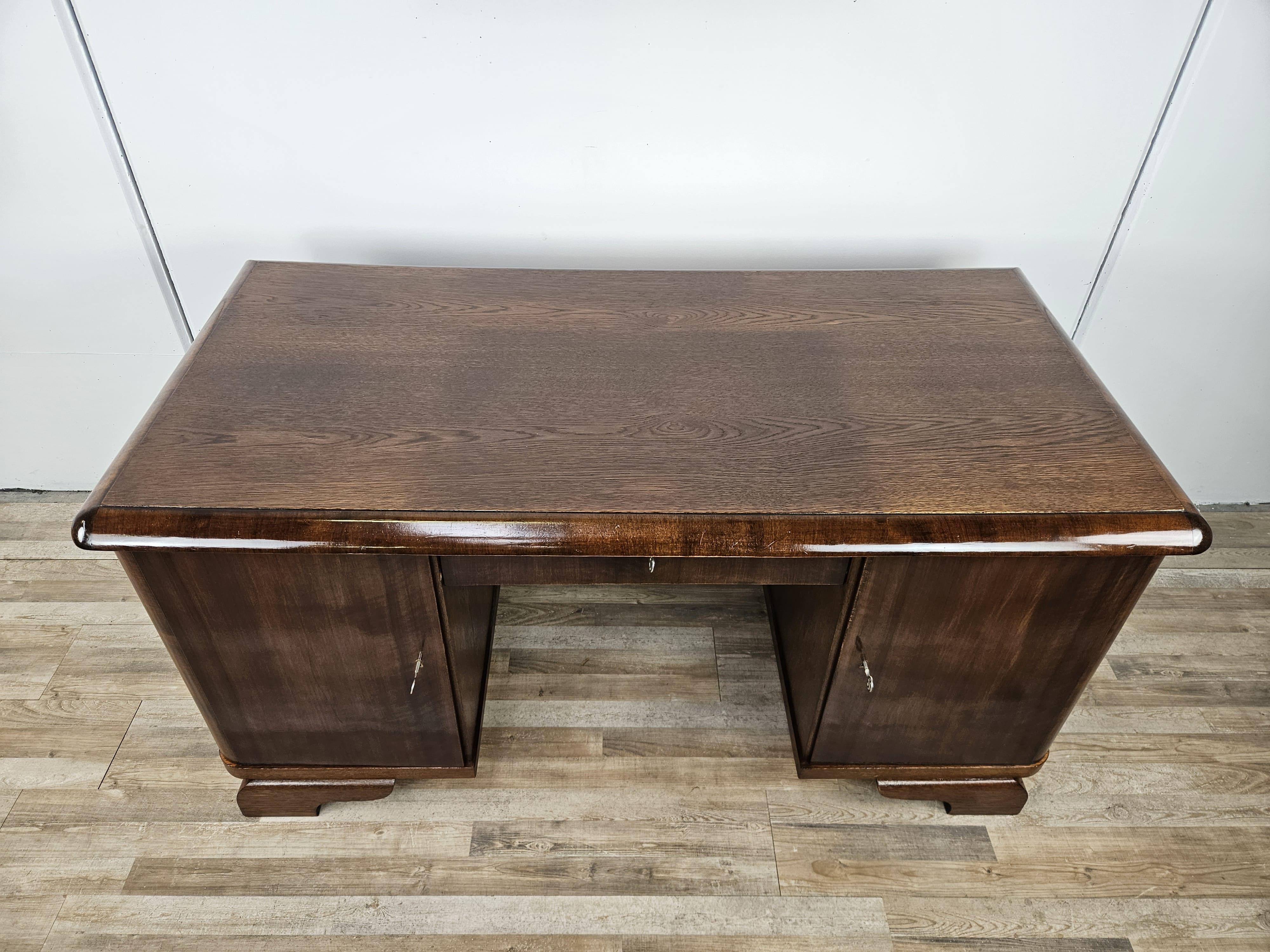 Elegante scrivania anni '30 di epoca Art Decò con struttura in legno di quercia, arricchita da due ante laterali e un cassetto centrale.

A sinistra troviamo tre ampi e robusti cassetti, sul lato destra due ripiani interni.
Al centro trova posto un