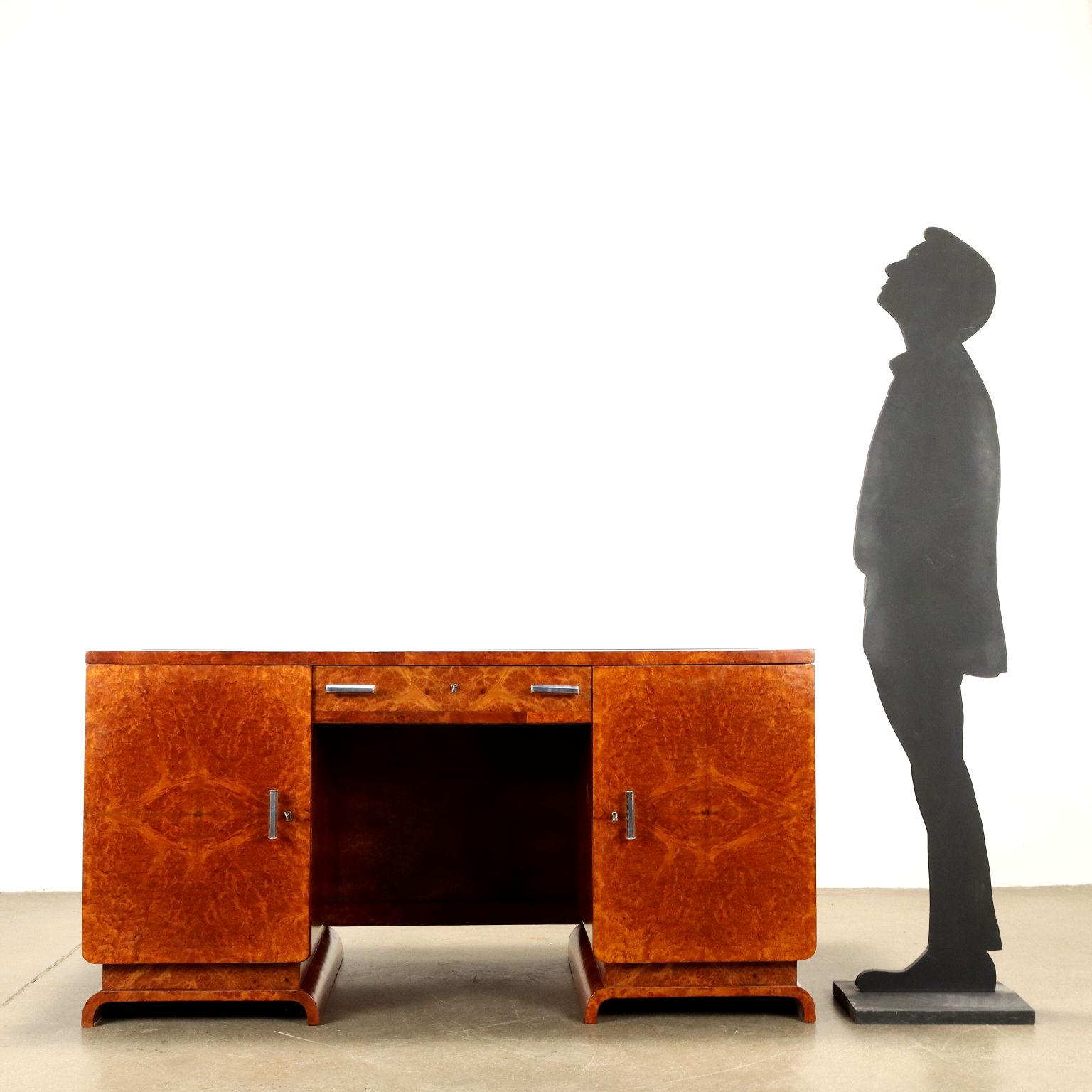 Elegante ed importante scrivania, con accurati e pregiati particolari costruttivi, vani laterali ad anta battente che contengono cassetti, cassetto sotto piano, maniglie in metallo cromato, legno impiallacciato radica di noce, piano in vetro