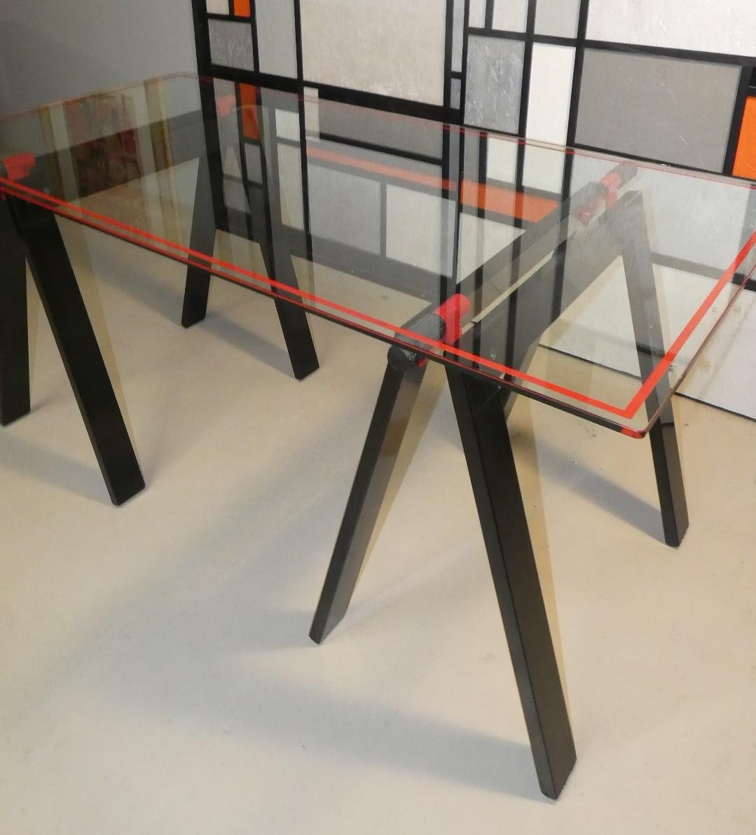 Mesa de trabajo Gaetano de Gae Aulenti para Zanotta. La encimera original de cristal biselado ribeteado en rojo, sostenida por dos caballetes plegables de aluminio esmaltado en negro con soportes de goma para los estantes. Un mueble que sigue