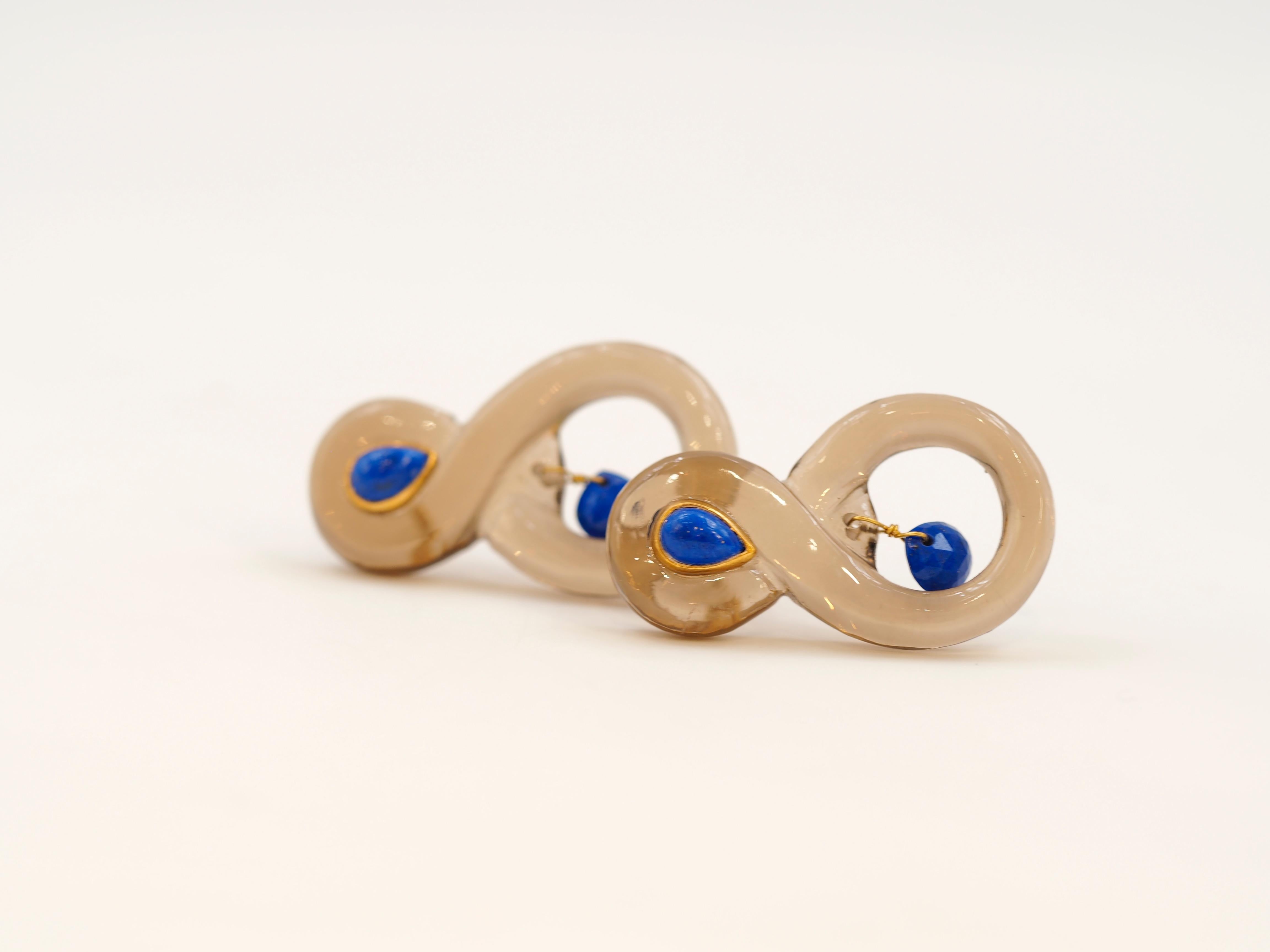 These earrings by Scrives are entirely hand carved smoky quartz and lapis lazuli. The main part has an 8 symmetrical shape to better adapt to the contour of the face. They are light on the ear (only 3.8gr). 
The stones can exhibit visible natural