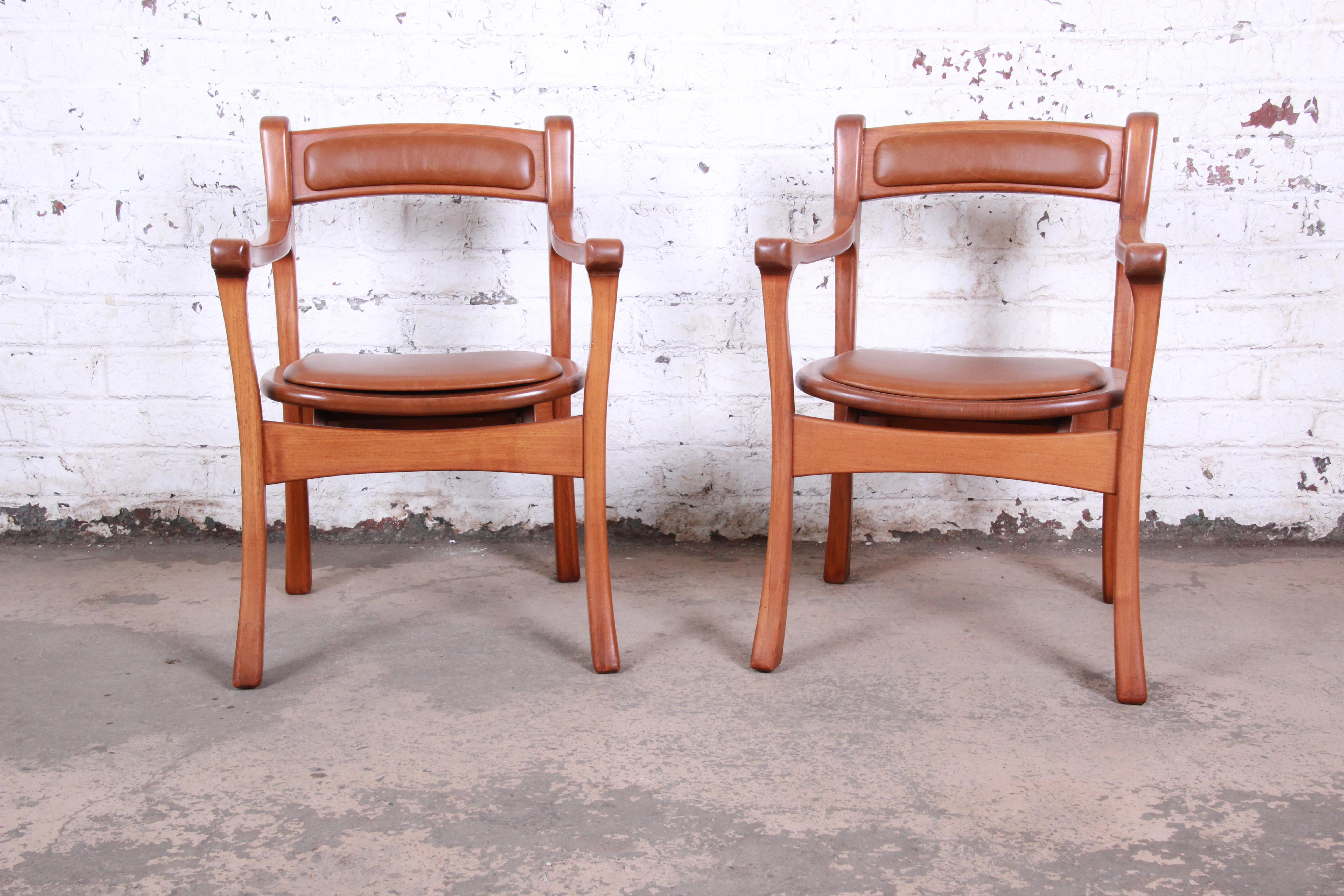 An outstanding pair of sculpted solid teak studio craft club chairs. The chairs feature unique sculpted teak frames with flared legs. They have been expertly reupholstered in brown leather. The chairs are in excellent condition. Ready for immediate