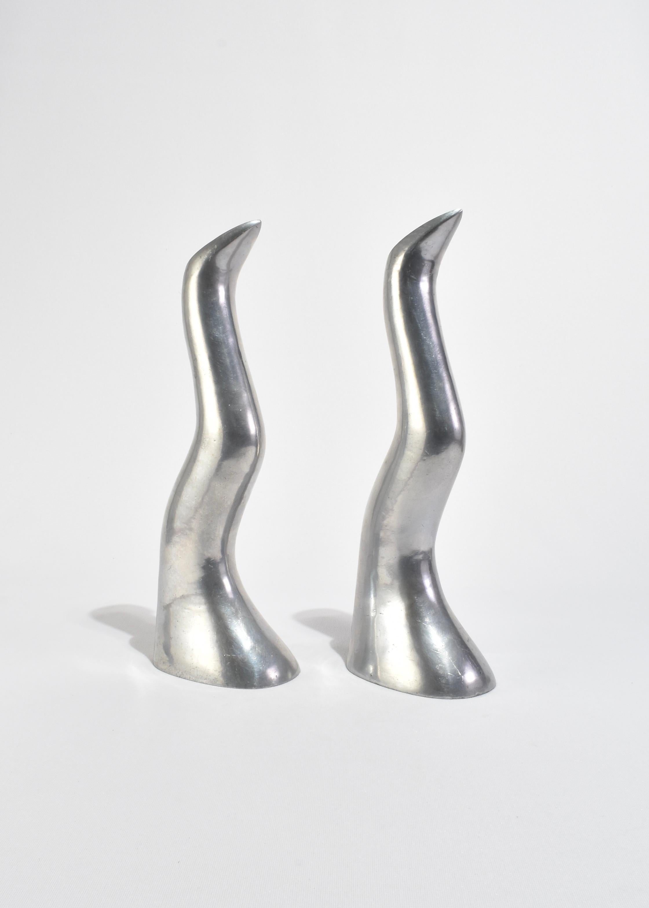 A pair of sculptural candlesticks cast in polished aluminum with fluid, tapering forms. Each piece rises in a graceful curve, ending in a narrow opening to hold a single taper candle. Their mirrored surfaces catch the light beautifully, making them