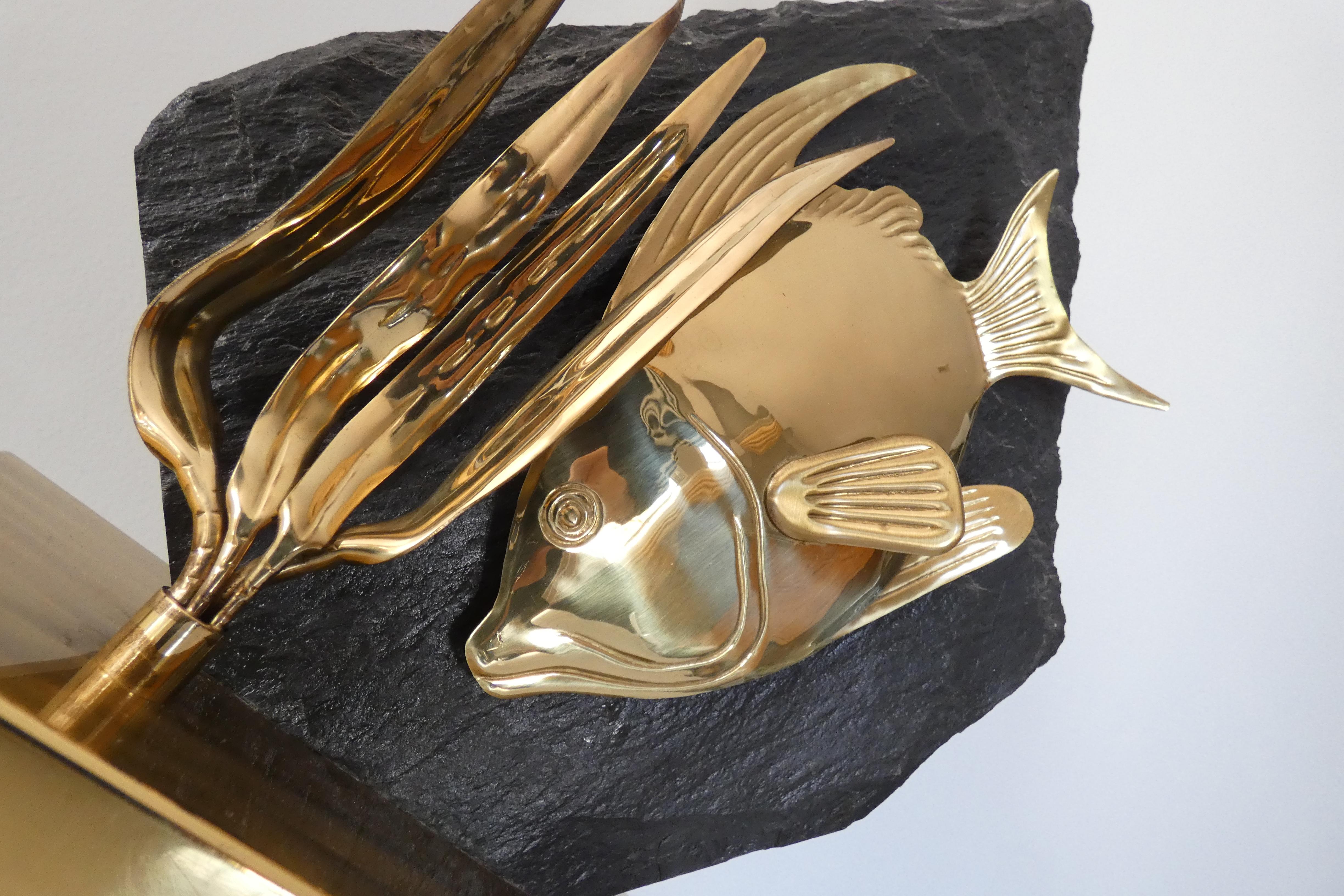 Lampe de table des années 1970. Cette pièce présente une sculpture en laiton poli représentant un poisson stylisé et des roseaux aquatiques, qui est montée sur une grande dalle brute de pierre naturelle foncée. Cette composition repose sur une base