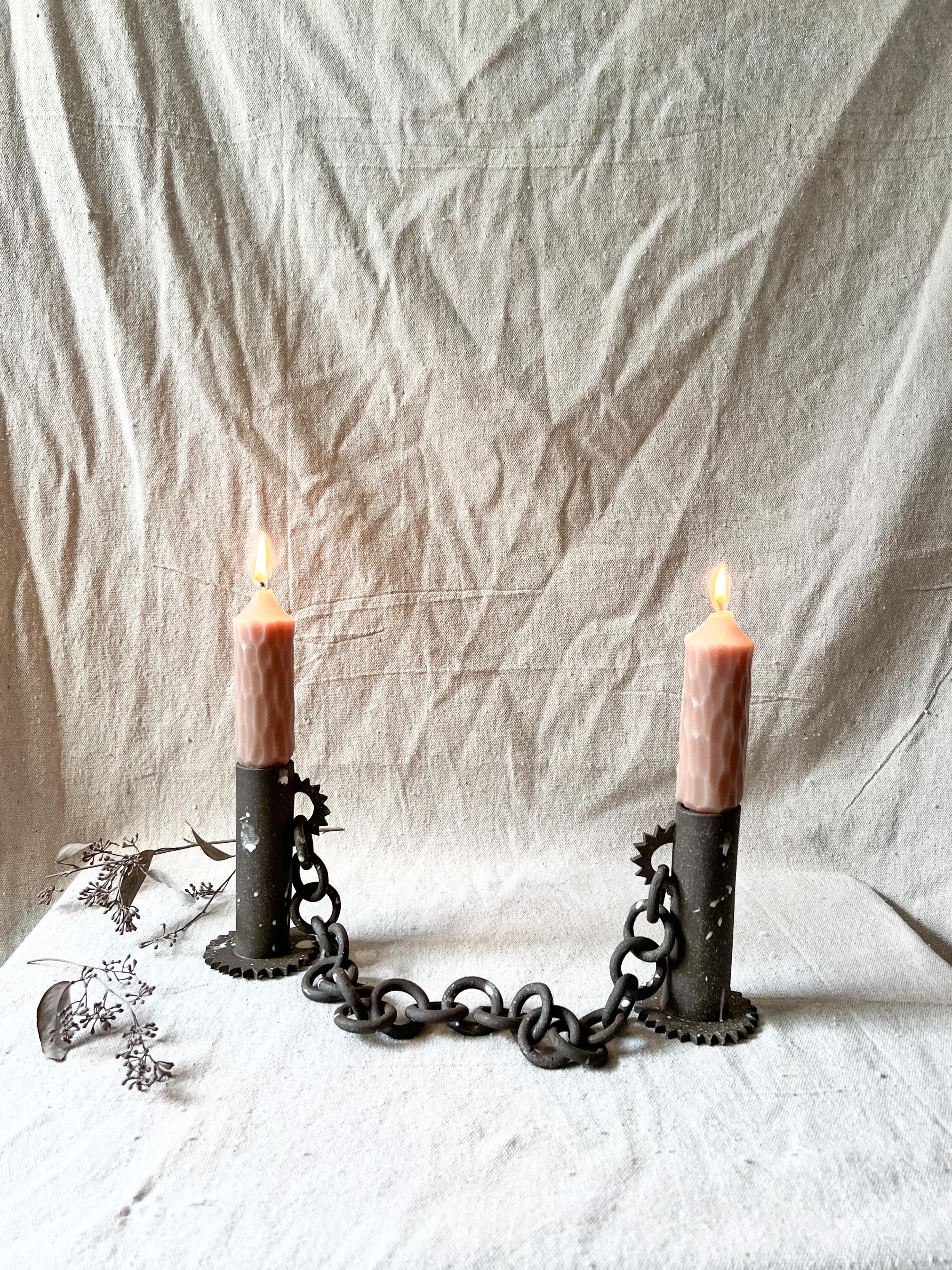 Deux bougeoirs sculpturaux en grès fabriqués à la main et reliés par une chaîne en céramique dégagent un charme éclectique, mêlant formes organiques et design fonctionnel. Ces bougeoirs uniques en leur genre présentent un caractère individuel et un