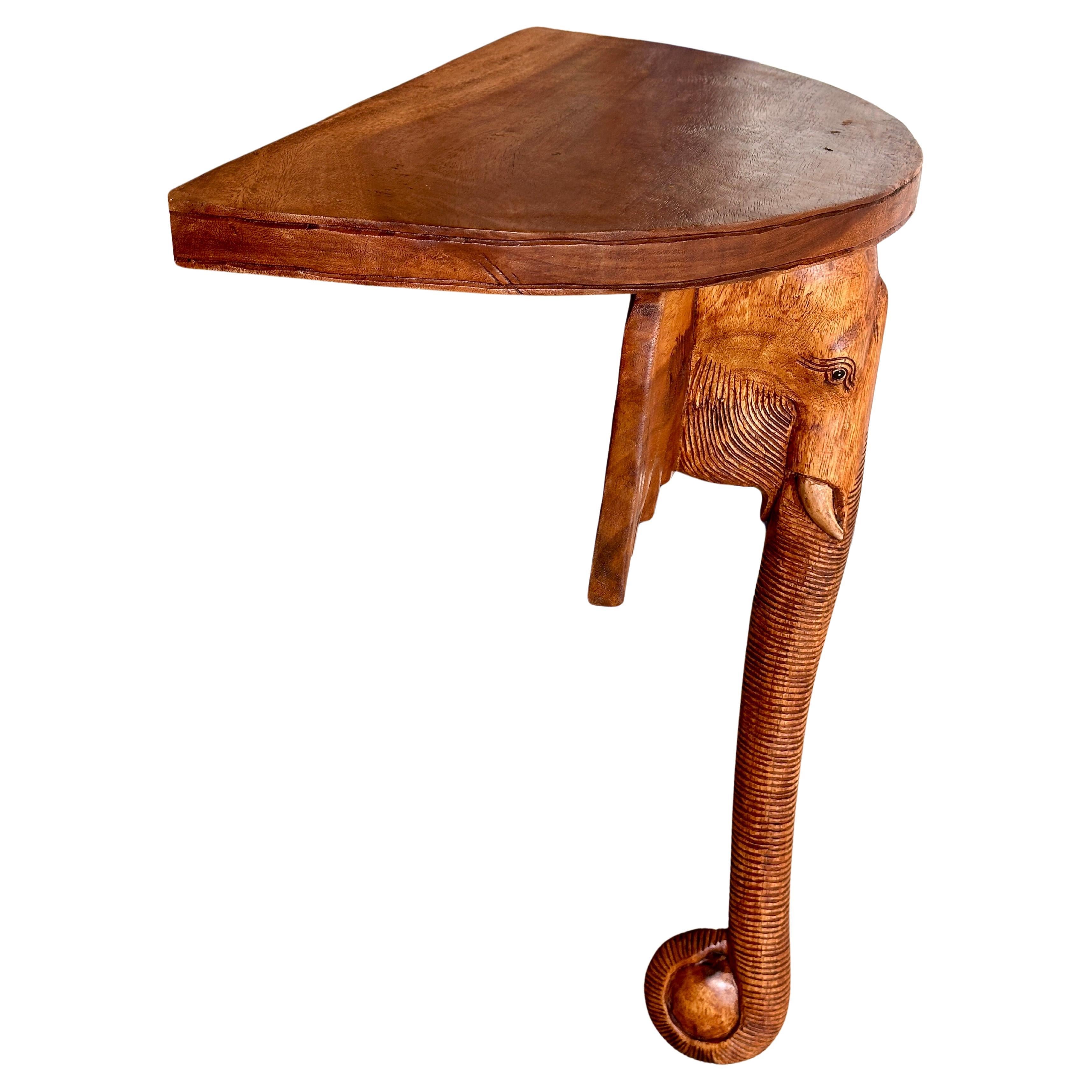 This striking demi-lune wall shelf features a beautifully carved elephant head with intricate details and a graceful trunk extending down as the main support. Crafted from rich, warm wood, the sculptural design adds an exotic and artistic touch to
