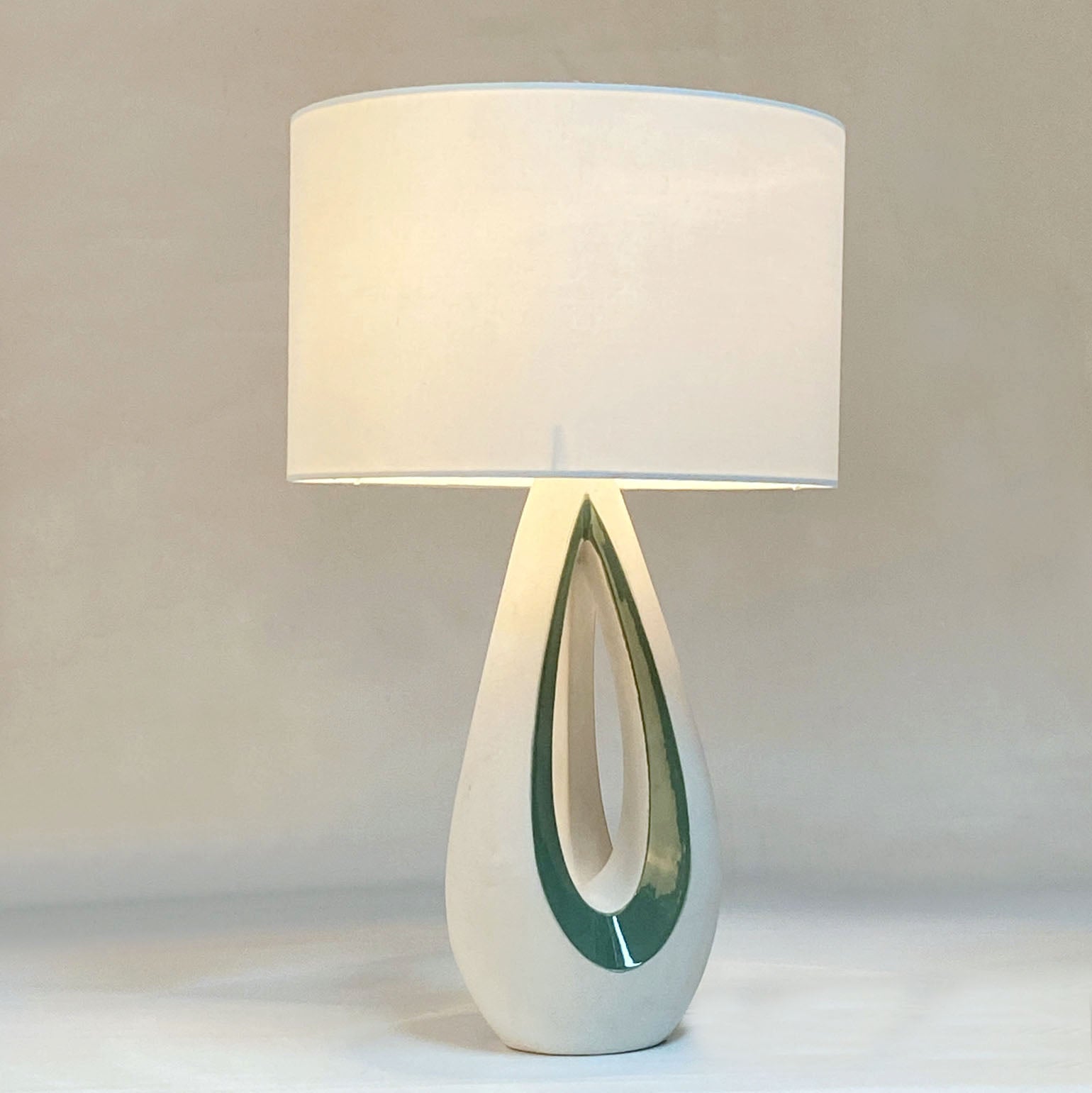 Elegant sculptural ceramic 1950's French table lamp in white and green glaze. The curves and openings in the shape of the base are accentuated by the green tear shape borders. The lamp comes with a cream drum lamp shade.

Dimensions Base; height 40