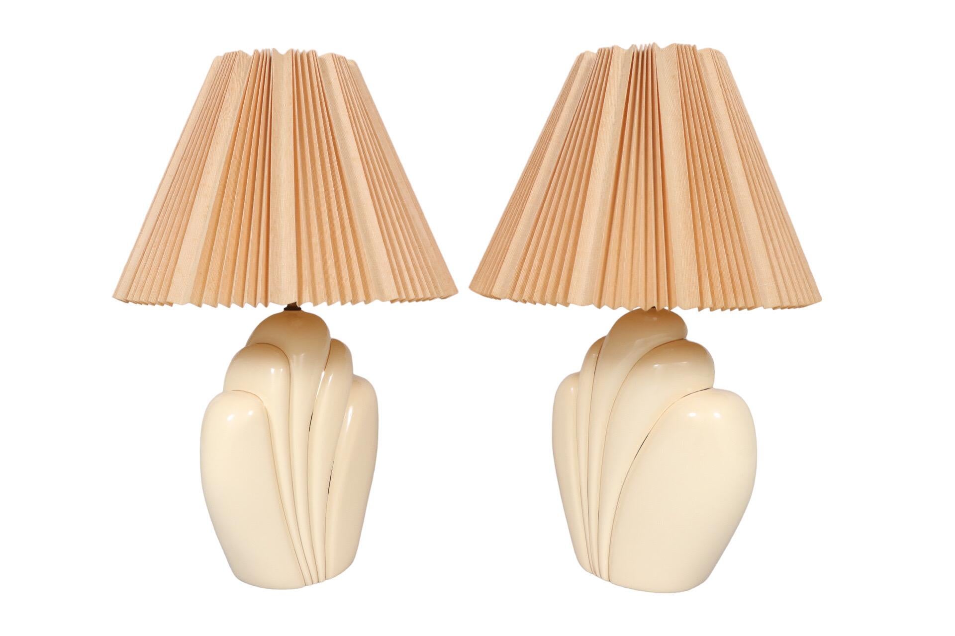 A pair of sculptural ceramic table lamps in cream. Vases are shaped like feather plumes trimmed with gilt lines. Lamps are topped with pleated empire shades in peach, topped with wooden ball finials. Wired and working. Each lamp measures 14