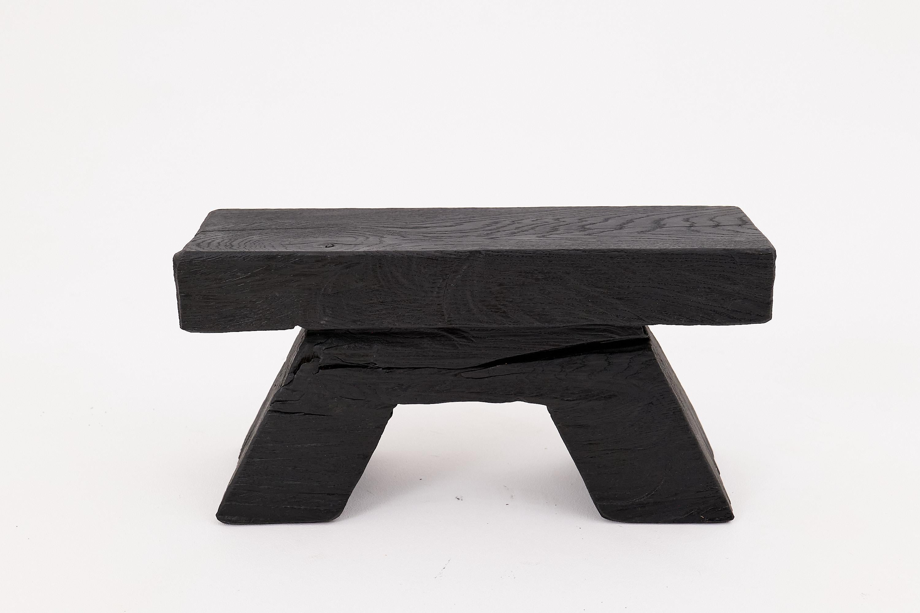 This bold charred wood stool blends elemental strength with modern geometric refinement. Hand-carved from solid reclaimed hardwood, it features a weighty, monolithic top resting on two angular, trapezoidal legs, evoking the form of ancient stone