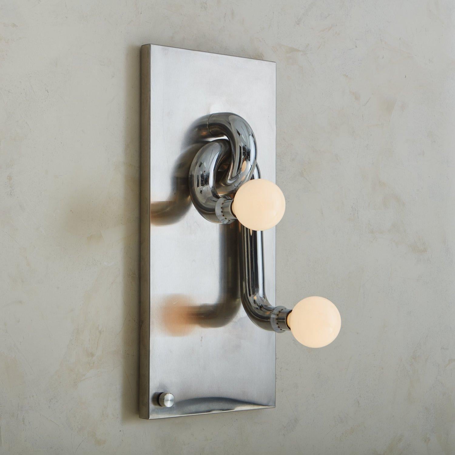 A sculptural Italian wall sconce with a rectangular chrome backplate, from which two intertwined, cylindrical arms extend. Each arm curves outwards at different lengths and supports one bulb. It turns on with a small circular knob on the lower left