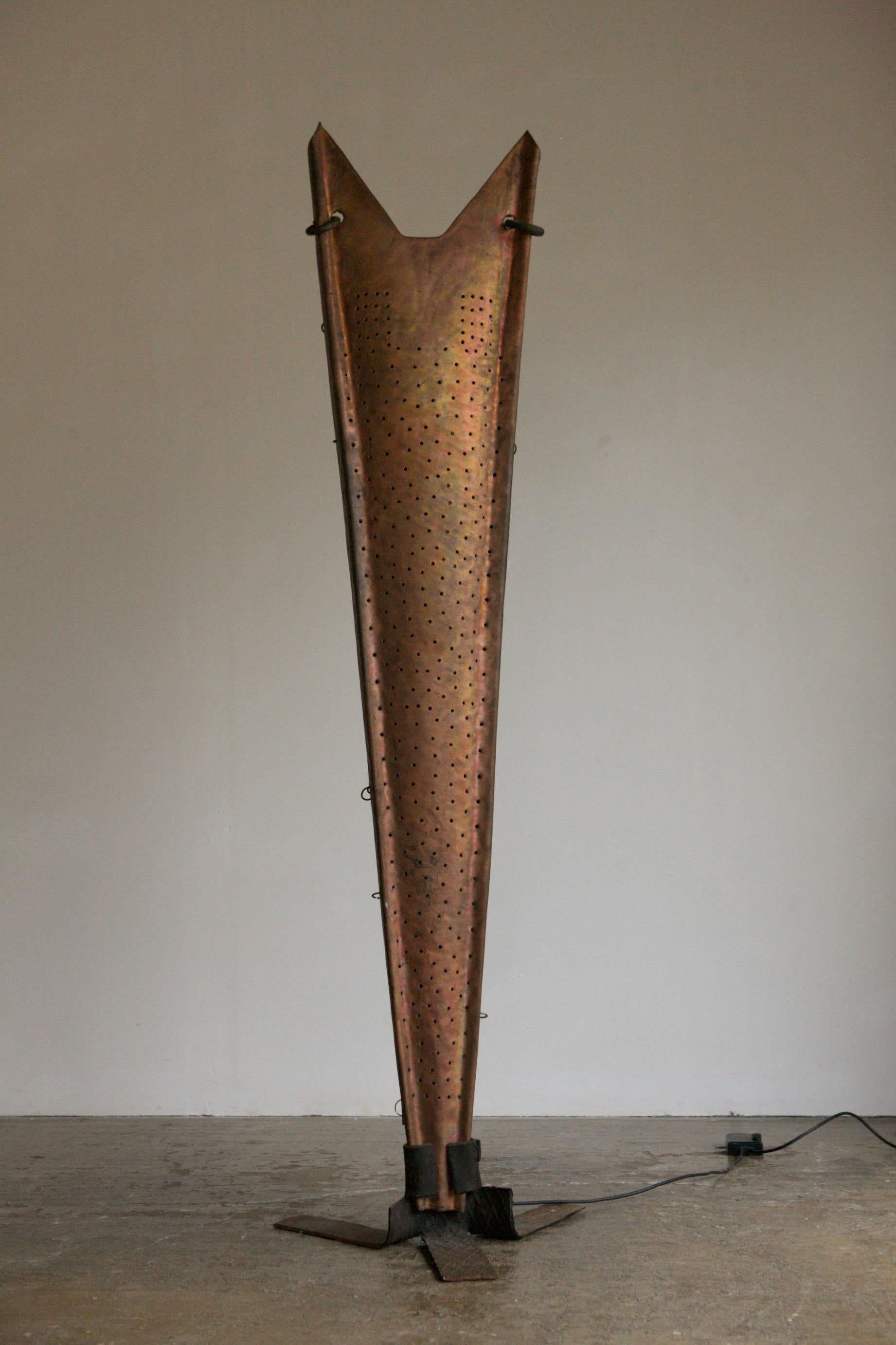 An outstanding floor lamp in hammered and pierced metal. This sculptural piece takes its design notes from the European brutalism. The shield like body is finished in a copper tone with metal hoops and gives off an up and back lit glow. 
