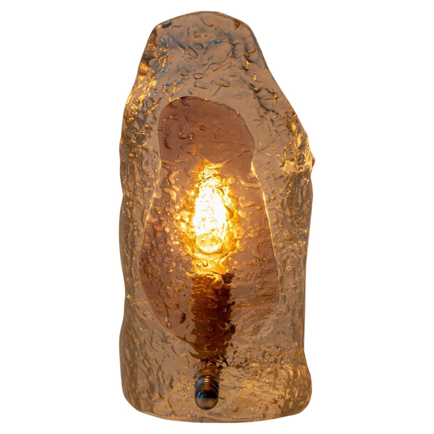 Sculptural mid-century table lamp in thick hand-blown lead glass. The organically tooled form combines clear glass with a soft mauve layer, its irregular surface alive with bubbles and texture that catch and diffuse the light. In full working order.