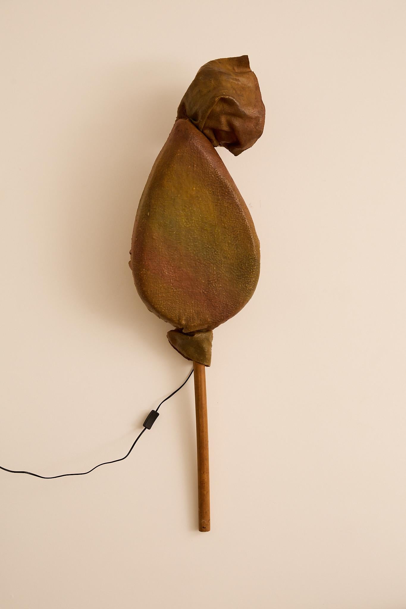 Lámpara escultórica de pie o de pared que se asemeja a un capullo envuelto, con una pantalla de fibra de vidrio o tela moldeada a mano en tonos terrosos, montada sobre un sencillo poste de madera. Cuando está encendida, la carcasa texturizada brilla