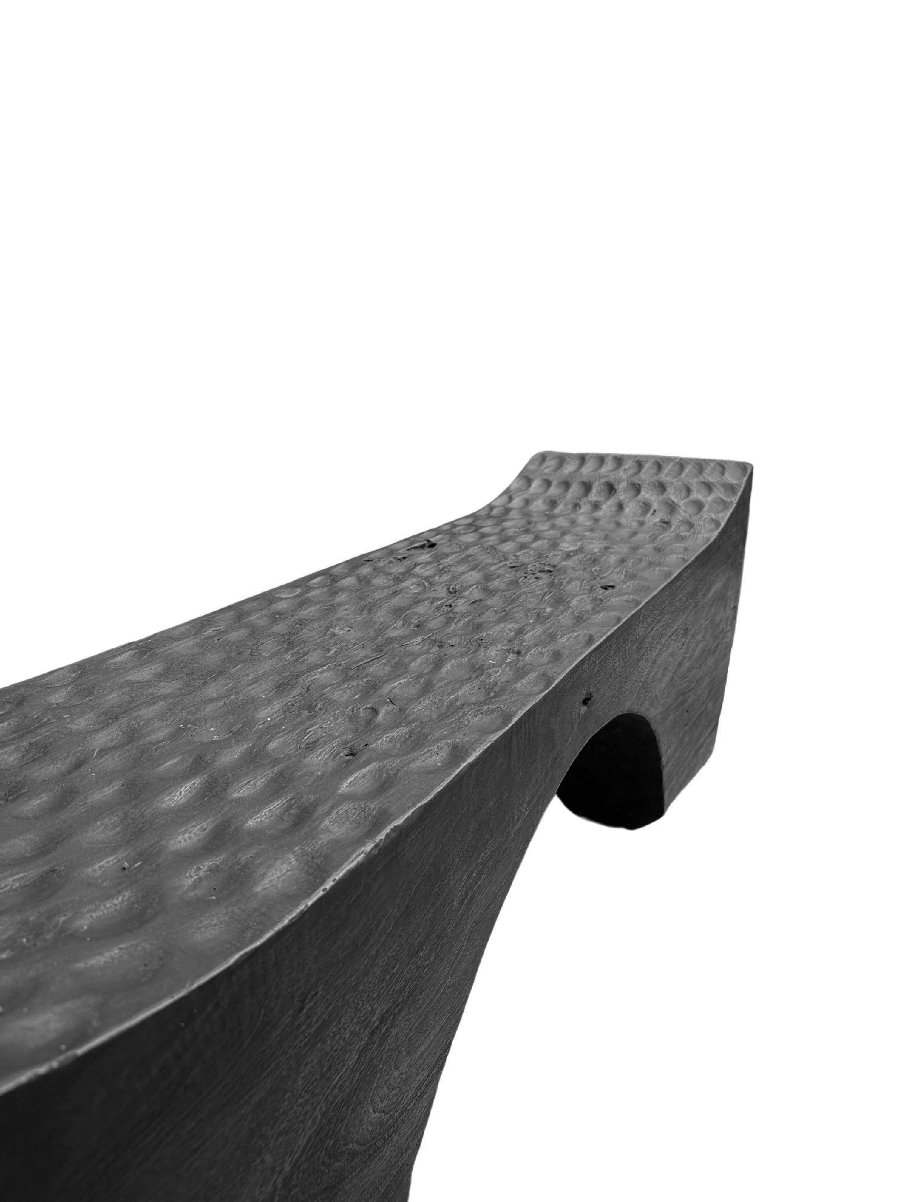 A sculptural hand-carved mango wood bench. To achieve is rich black pigment the bench was burnt three times and then finished with a clear coat. The mix of curved and straight lines along with a myriad of textures make for a very elegant bench.