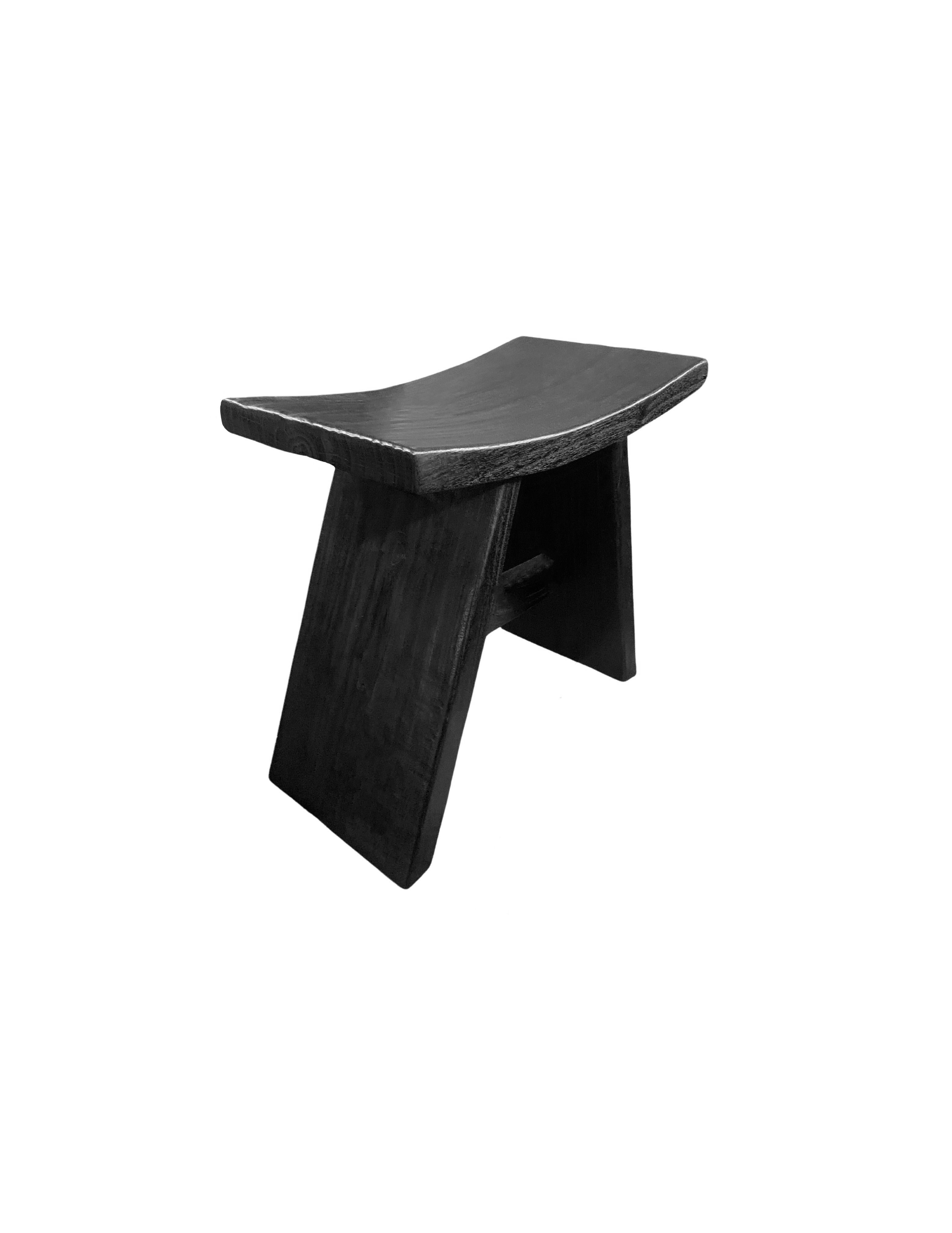 A wonderfully sculptural mango wood stool with angular legs and a curved seat. To achieve its rich black pigment the wood was burnt multiple times and then finished with a clear coat. Its neutral color and minimalist shape make it suitable for any