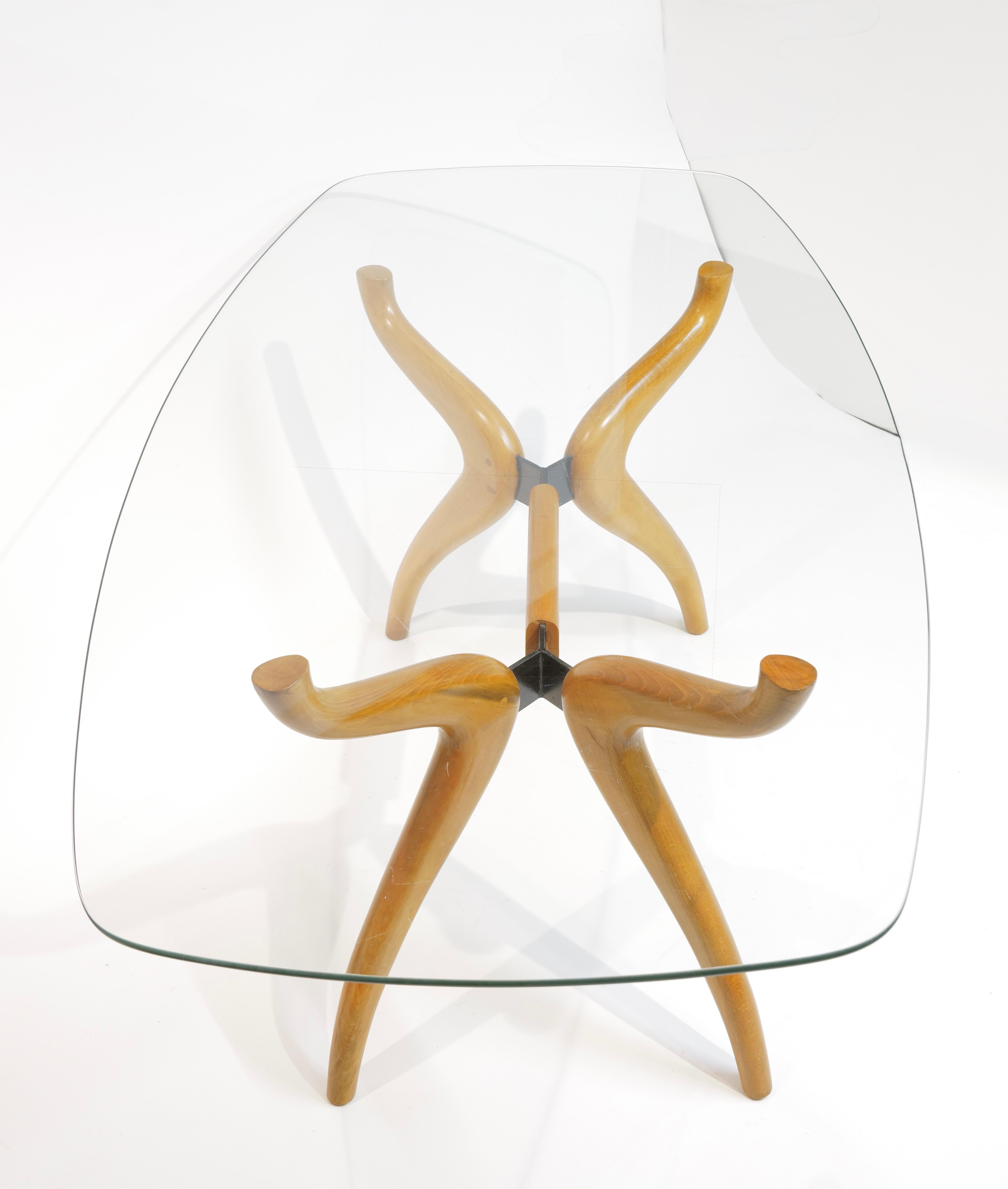 Llamativa mesa de comedor moderna de mediados de siglo, con una base de madera maciza esculpida con gracia y un tablero de cristal de forma orgánica. Las patas onduladas, elaboradas en cálido arce o haya, se cruzan en una junta central, creando una