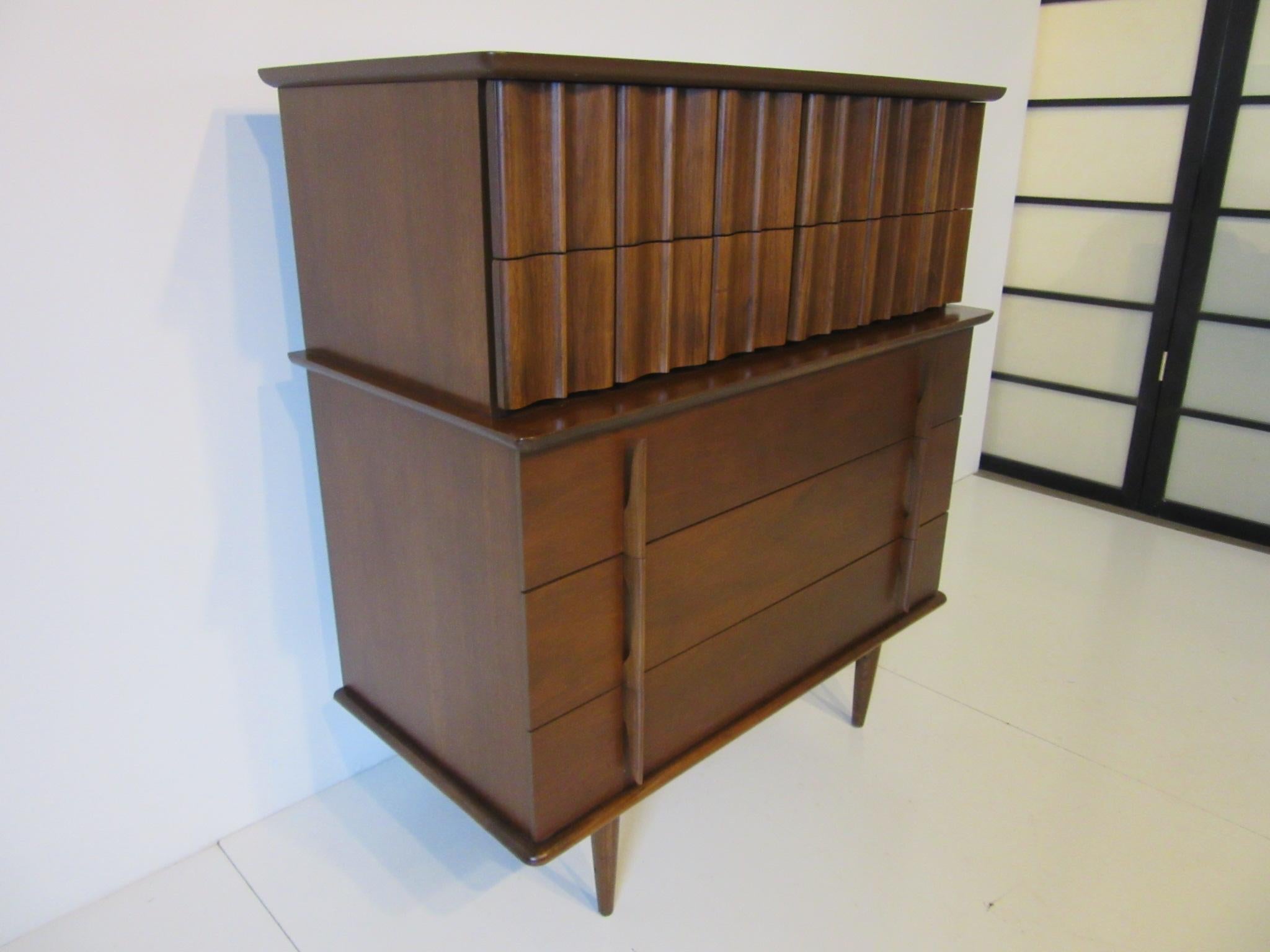 A dark walnut toned tall dresser with great sculptural handles and drawer fronts, an interesting take on an old chest on chest design transferred to a midcentury vibe with plenty of storage.