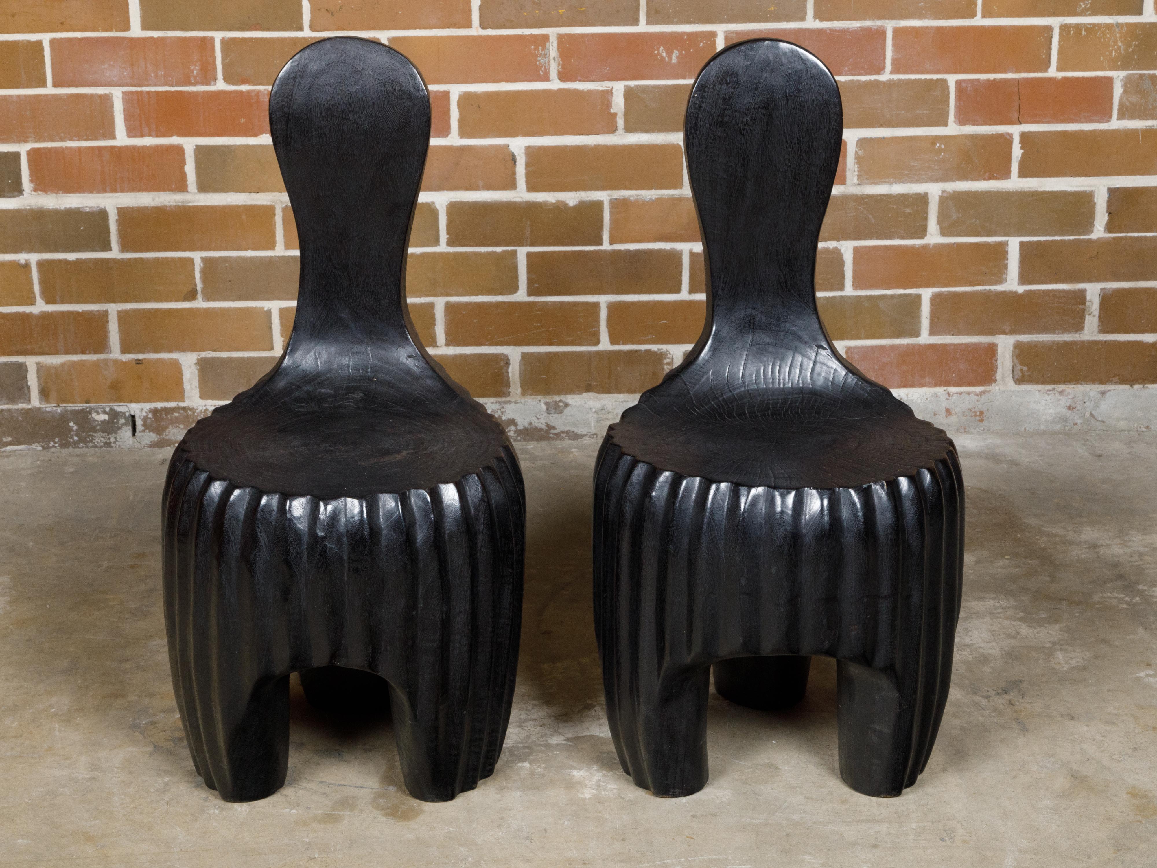 A sculptural pair of Midcentury African wood spoon back black chairs with carved reeded bases. This sculptural pair of Midcentury African wood chairs embodies the essence of artistic furniture design. With their distinctive spoon back silhouette,