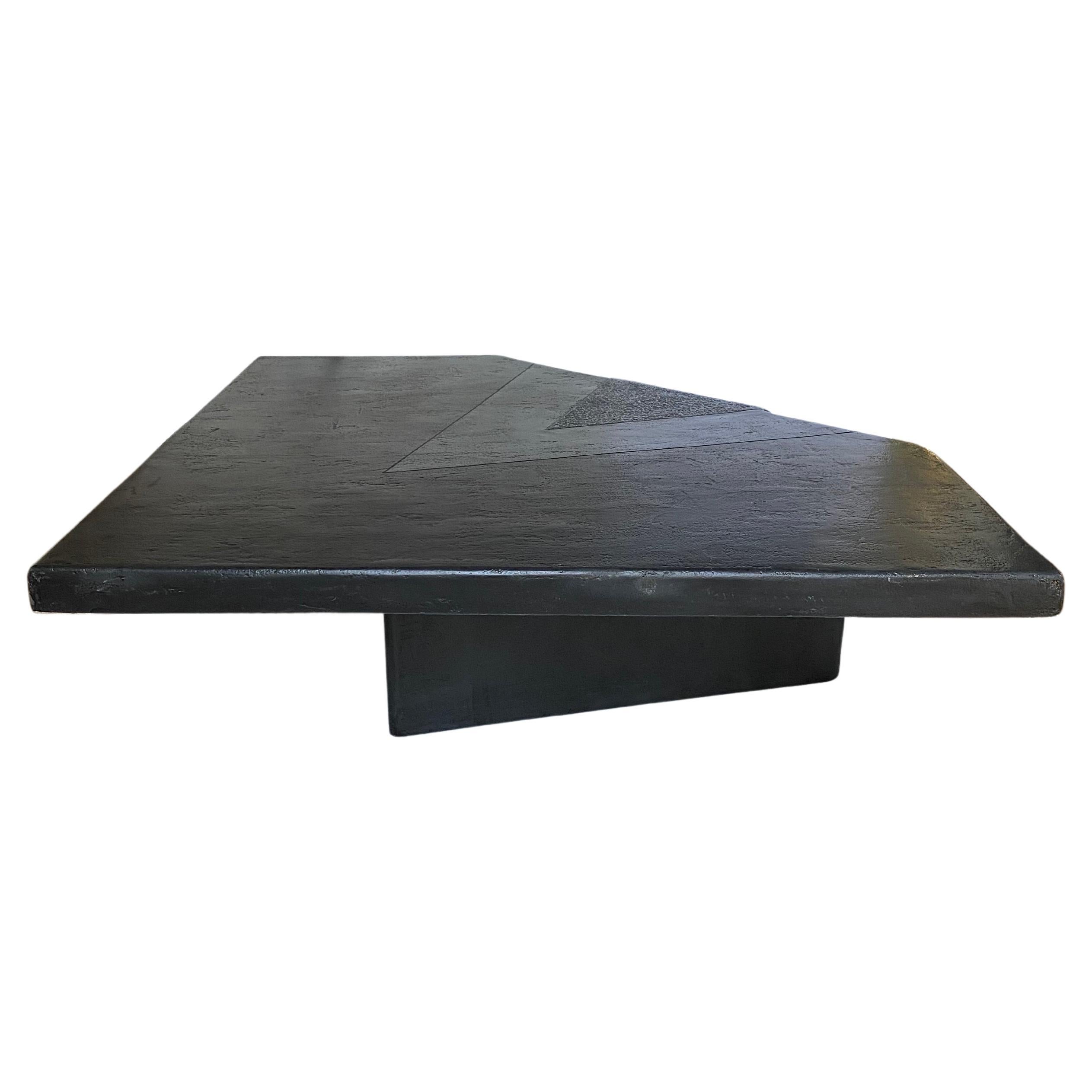 Sleek and sculptural black plaster coffee or cocktail table. Features a unique geometric, polygon, triangular shaped top on a cantilevered base with dramatic waterfall effect on one corner of table. Colors are black, grey and charcoal. The table’s