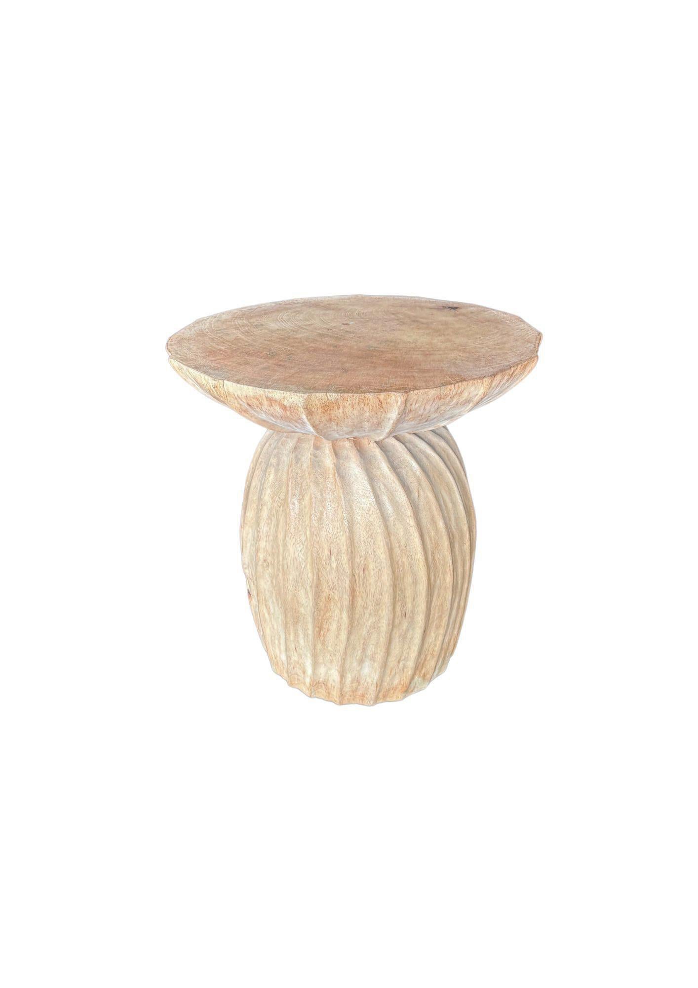 This mango wood side table features a beautifully sculpted design with a unique bleached finish, giving it a soft, washed-out tone. The natural grain of the mango wood is still visible, adding texture and character to the piece. The table's base