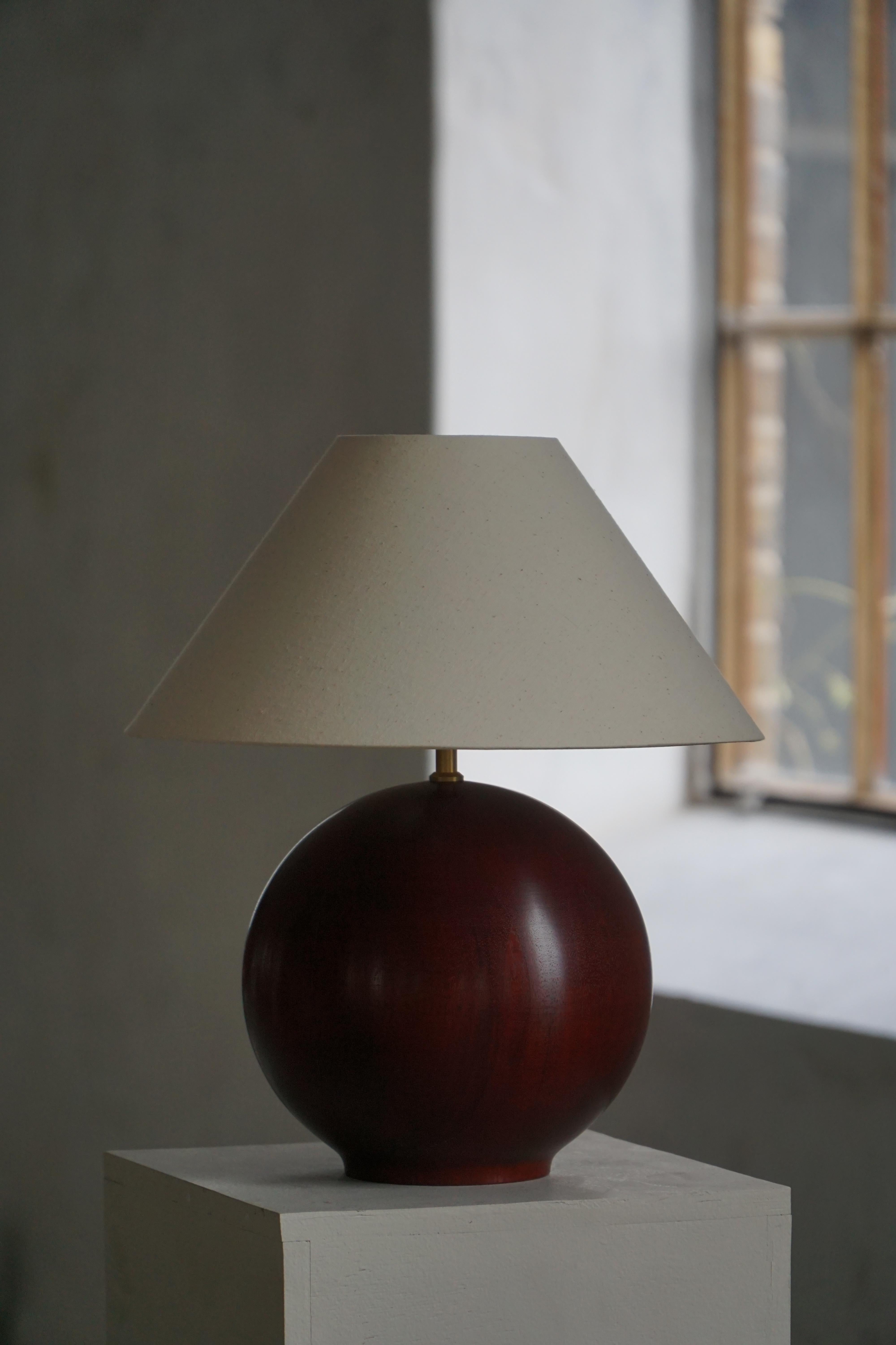 A sculptural table lamp crafted in dark stained beech wood with a warm reddish gloss, featuring a smooth, sphere-shaped base that emphasizes its minimalist and modern form. The lamp’s rounded silhouette highlights gentle curves and the subtle grain