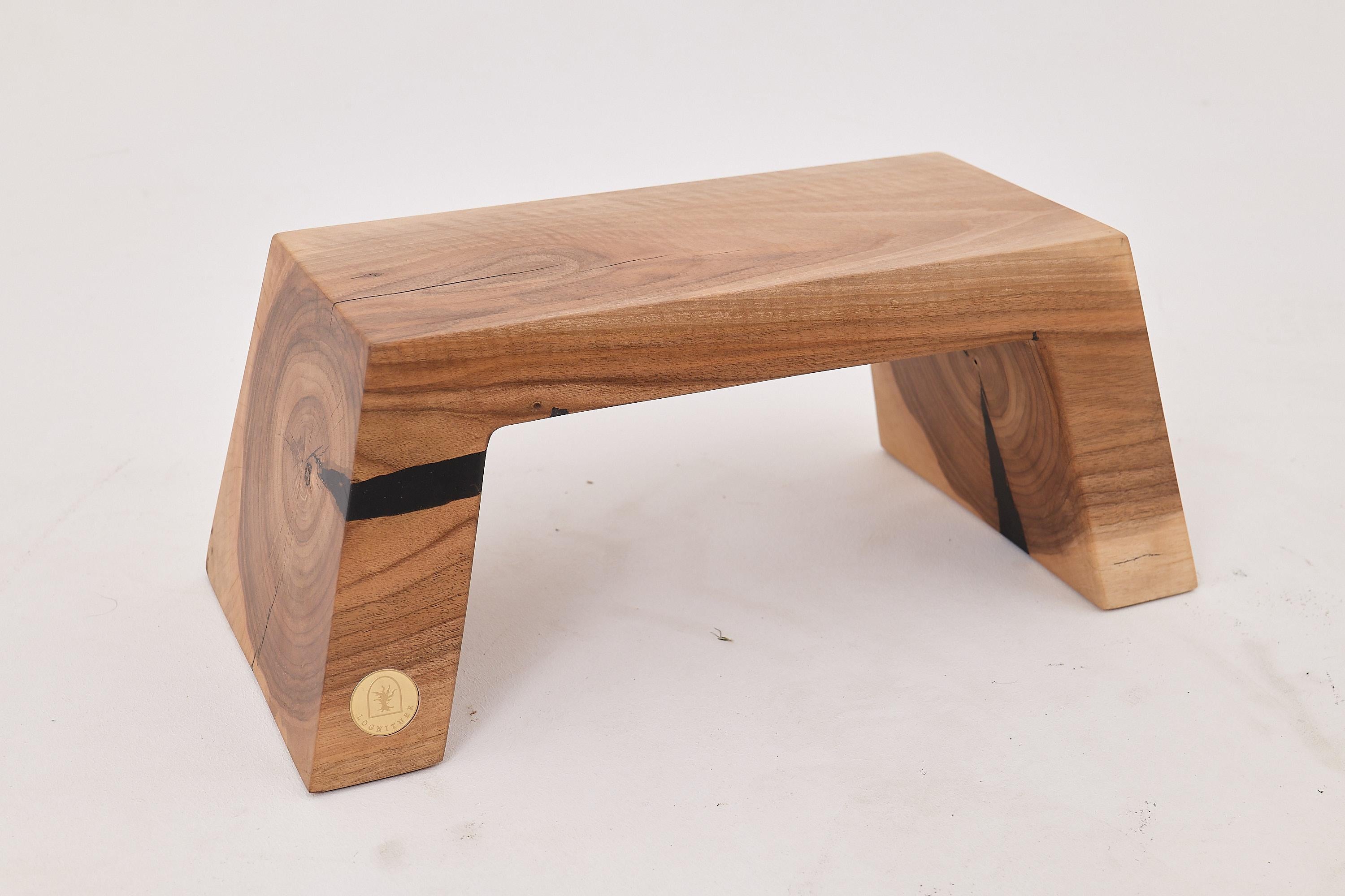 Este banco escultural es un estudio de pureza y contraste, tallado en una sola pieza de madera maciza y moldeado con una atrevida forma geométrica. Sus patas cónicas y su perfil monolítico se suavizan con la calidez del veteado natural, revelando