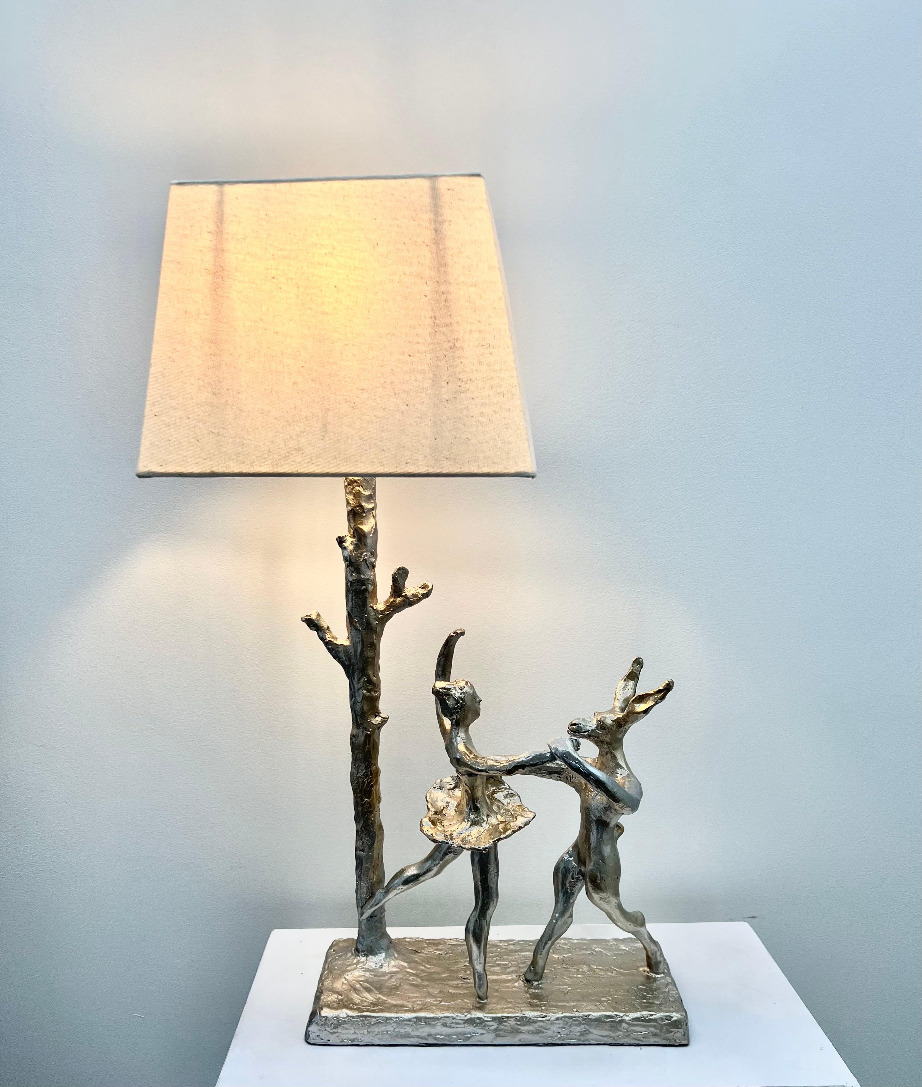 A  sculptural table lamp of a  hare gracefully dancing with a ballet dancer, posing by a tree trunk.  A charming functional artwork, hand crafted, moulded and cast in a lighter resin.  All parts are individually crafted, cast and assembled and each