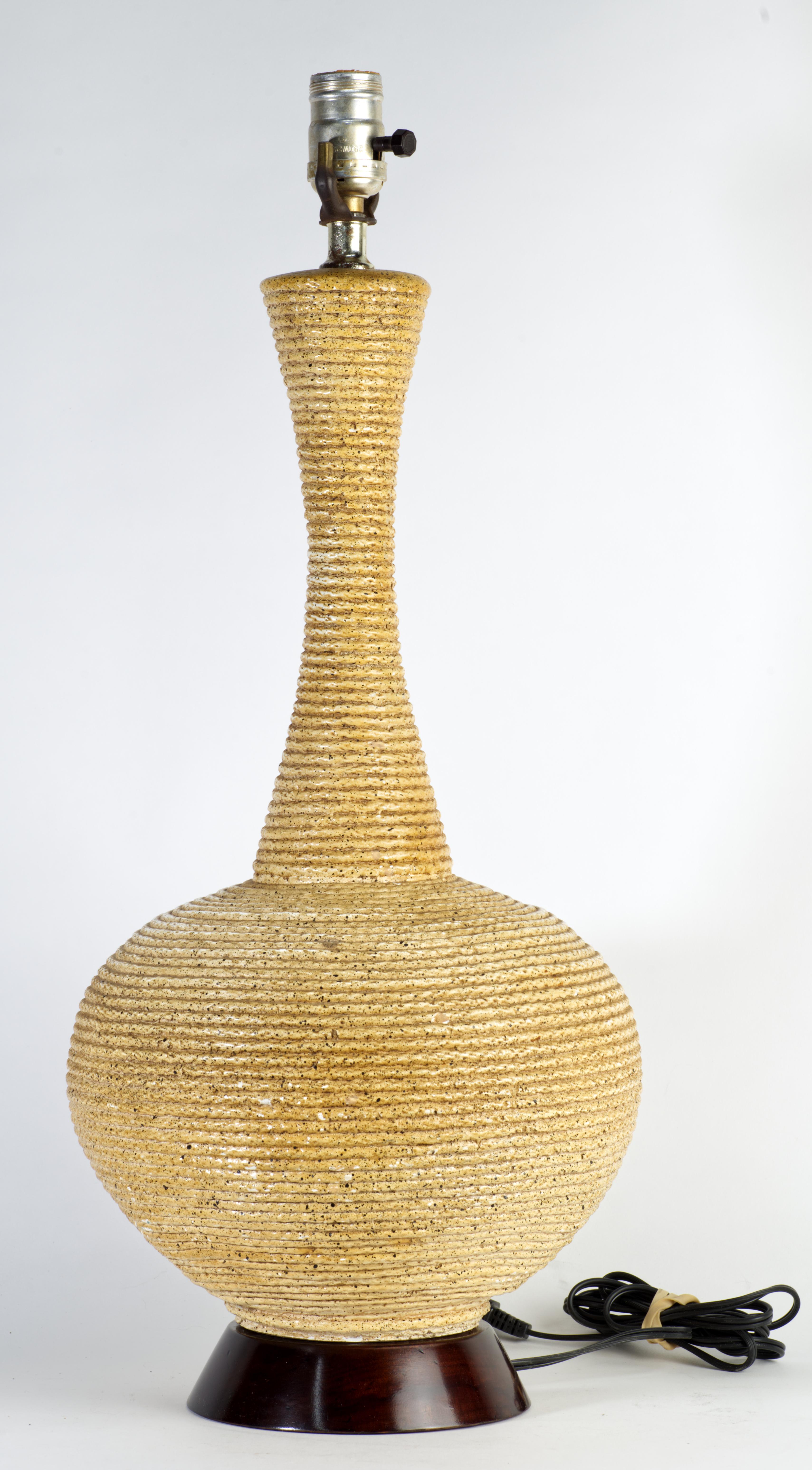 Cette lampe de table moderne inhabituelle est dotée d'un corps sculptural et graphique en céramique, dont la texture très détaillée rappelle la corde qui entoure l'ensemble de la lampe. Le glaçage beige-brun légèrement moucheté ajoute encore à