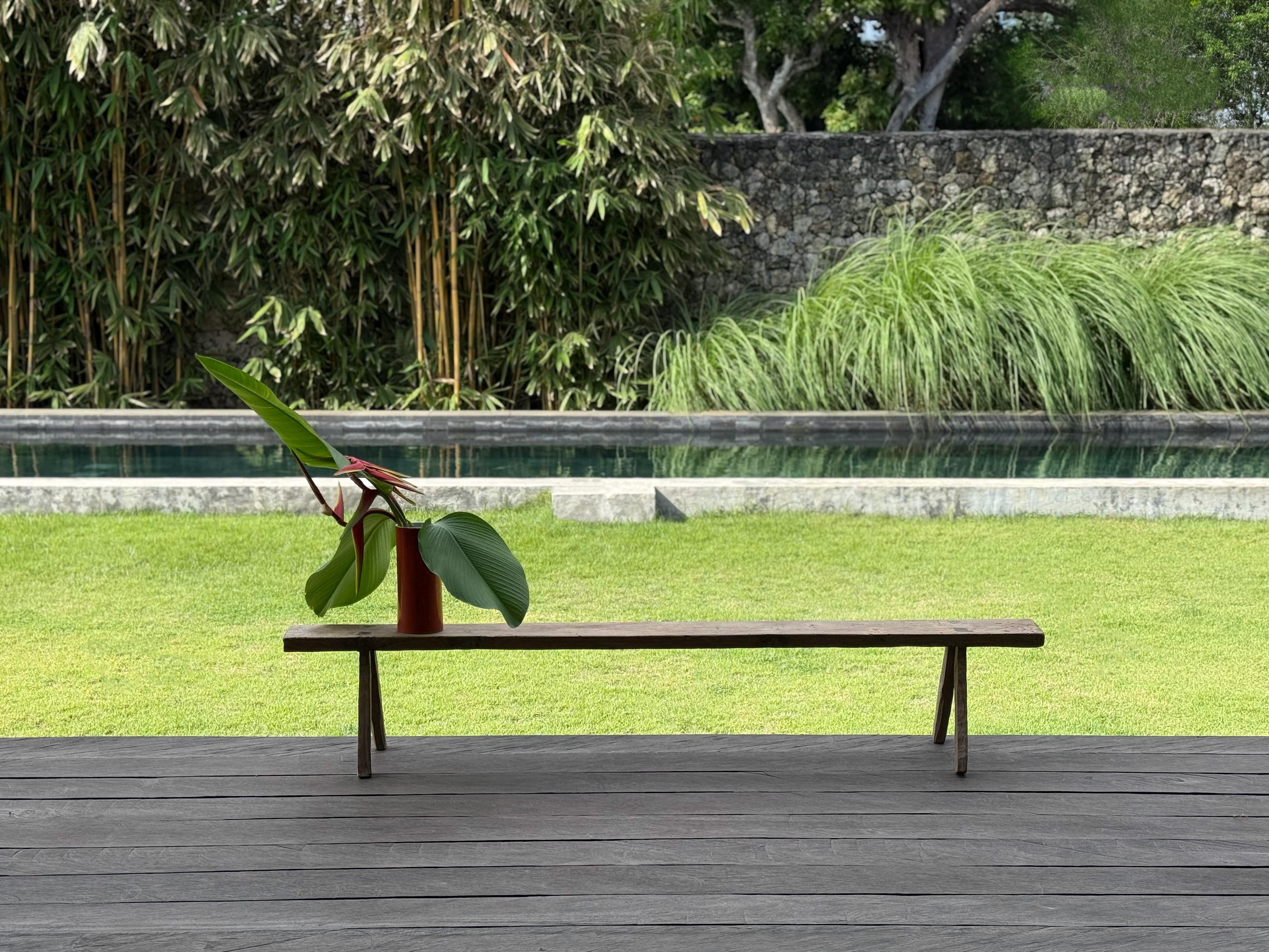 This hand-carved teak wood bench originates from the Island of Madura, off the coast of Northeastern Java. It features a long slender form with angular legs. A wonderful sculptural object with wood textures and shades. The seat was carved from a
