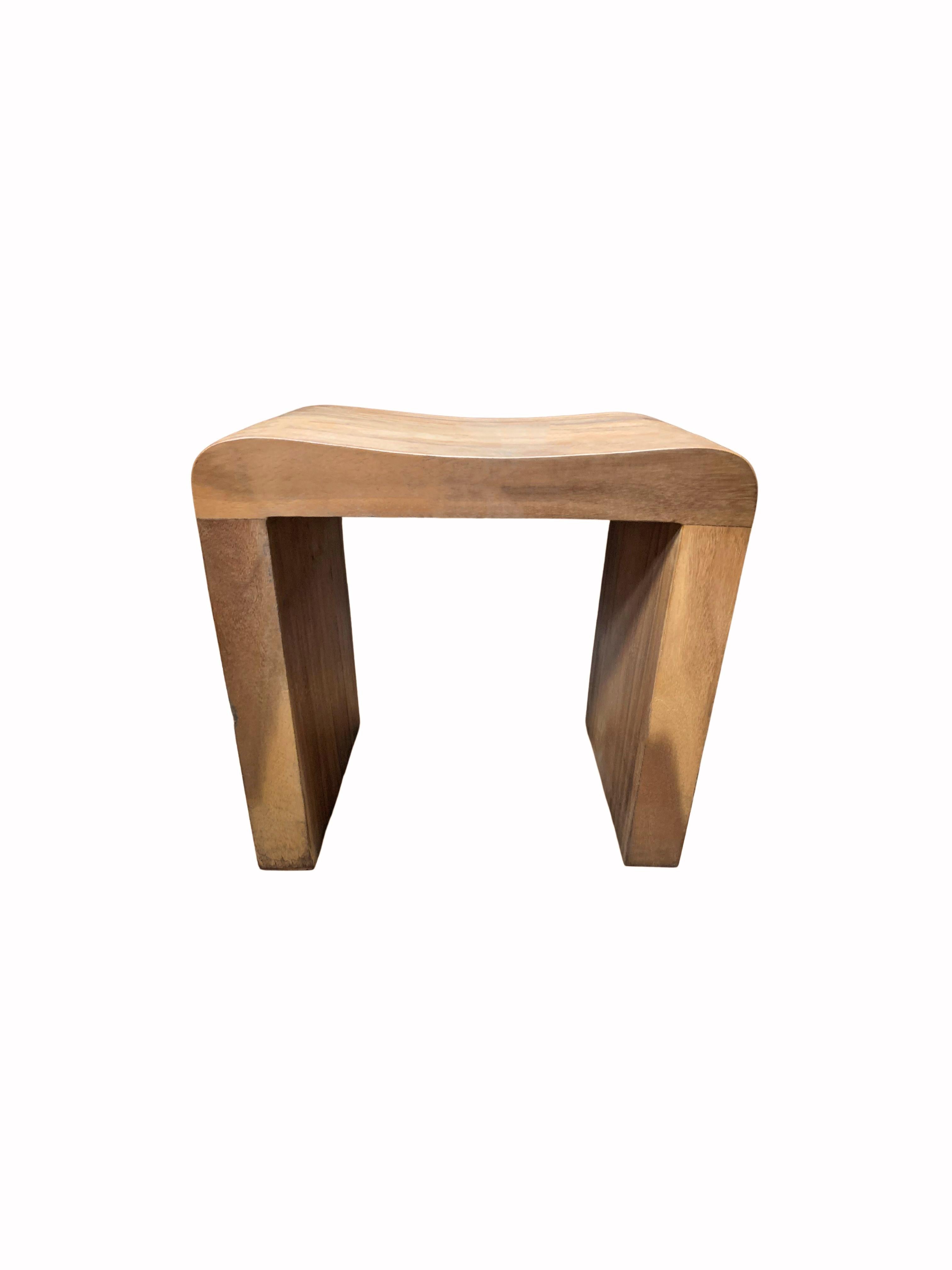 A wonderfully sculptural teak wood stool featuring a curved seat. The mix of wood textures and shades adds to its charm. Its neutral color and minimalist shape make it suitable for any space.