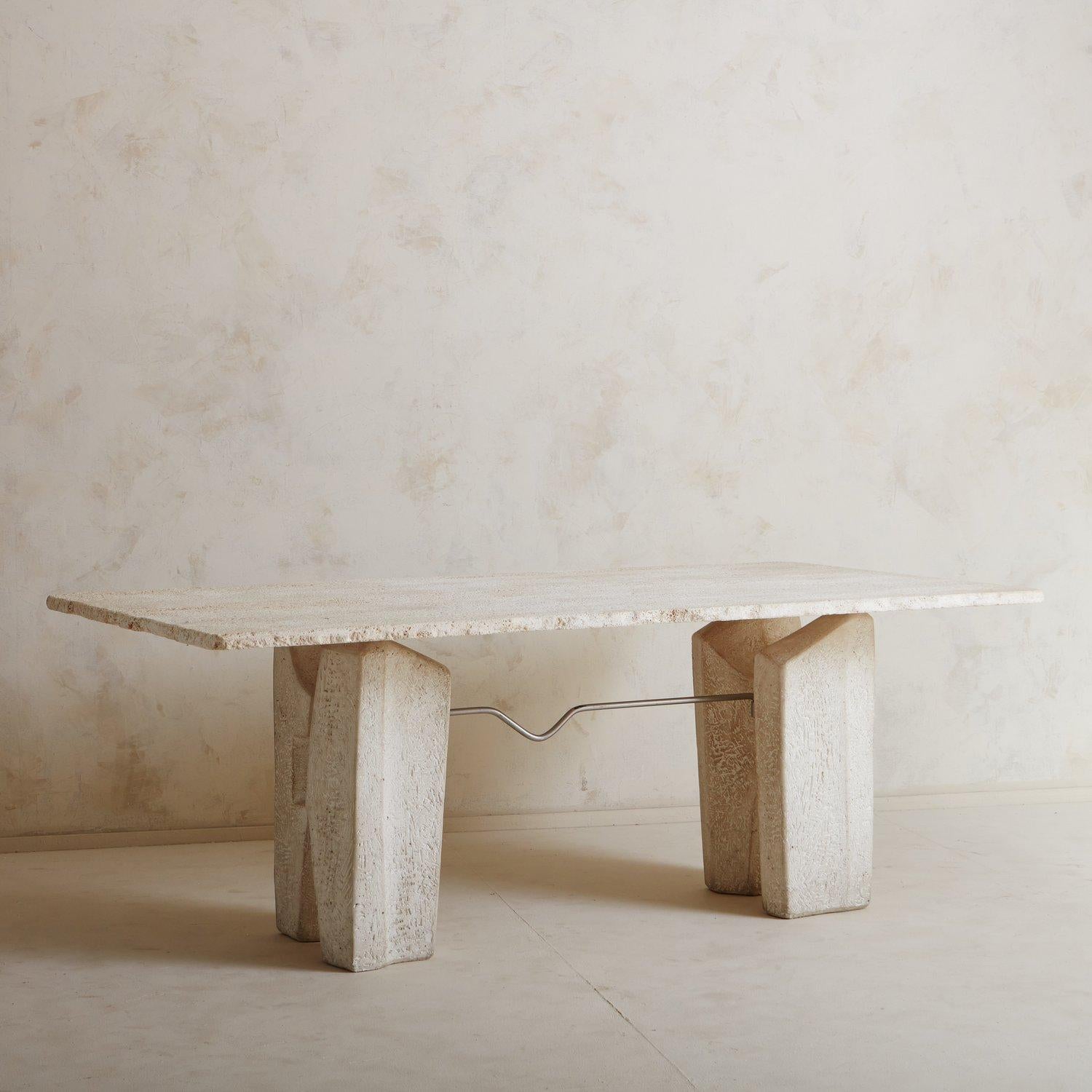 A stately 1970s dining table constructed with travertine. This piece features two sculptural trestle legs which support a thick rectangular top with raw, textured edges. The top sits just above the legs on metal supports, making it appear to float.