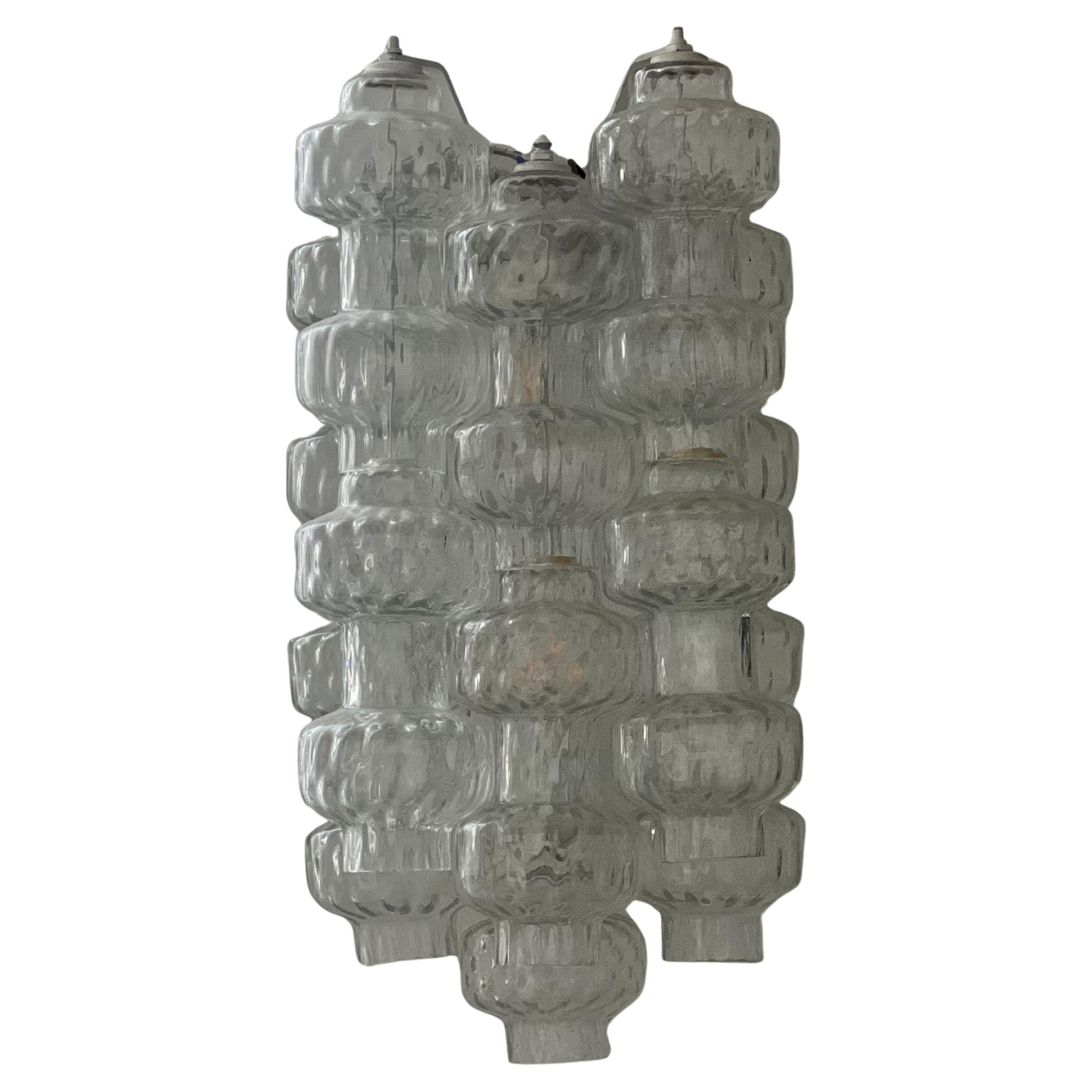 Aplique escultórico formado por una estructura de metal pintado de la que cuelgan seis pilas de dos piezas de cristal de Murano soplado. Una fuente de luz por aplique. redondeado y suave en la forma , crea un gran reflejo con la luz y parece una