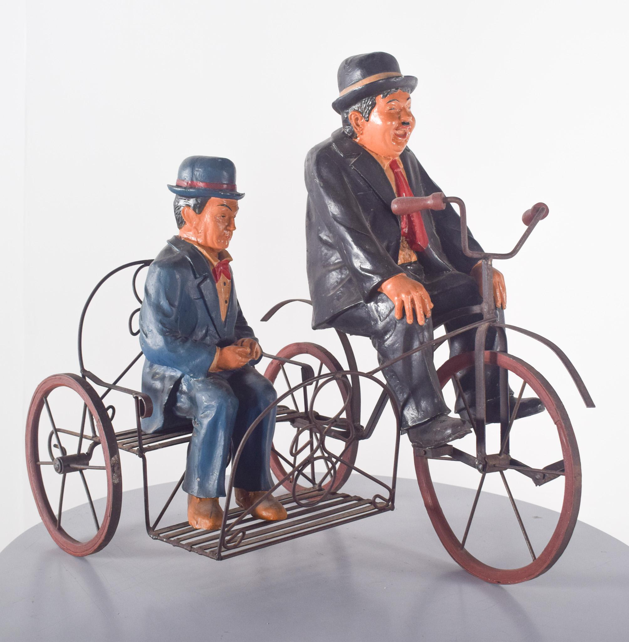 Sculpture depicting Bucha and Estica on a bicycle. The piece combines metal and painted resin, bringing the figures to life with caricatured expressions and formal attire consisting of tails, ties and short-brimmed hats.
The burly man in front,