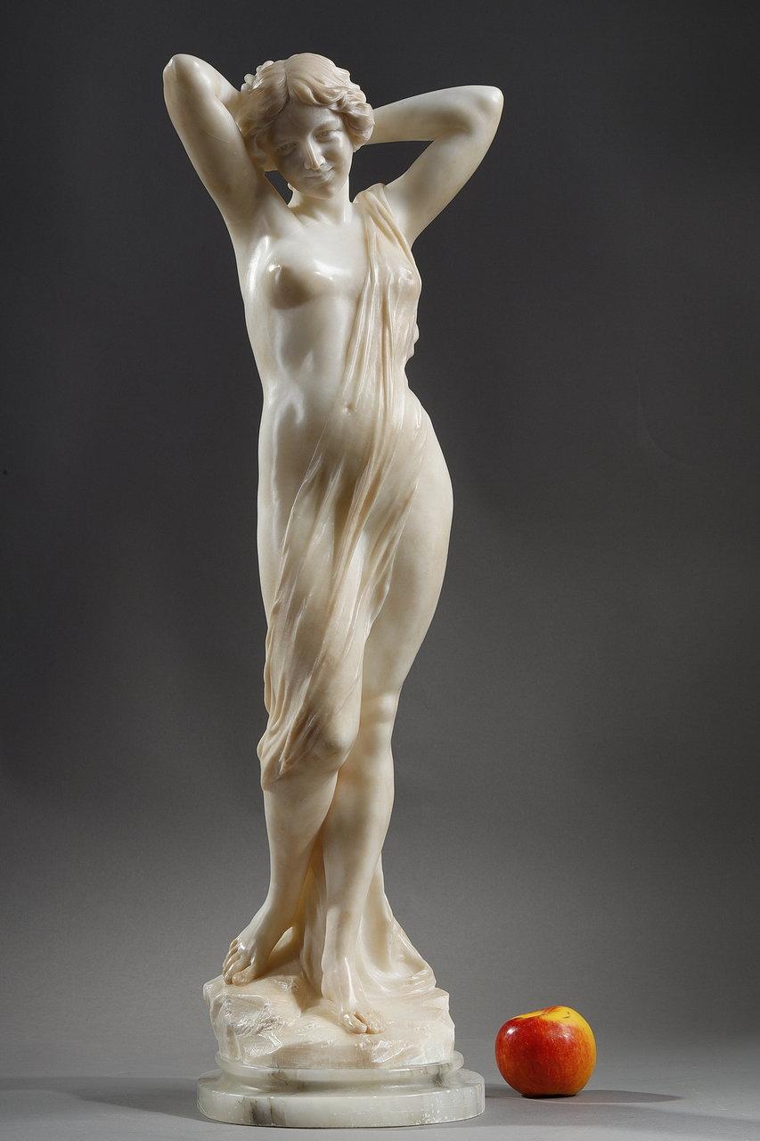 Italian sculpture in alabaster representing a young naked woman in the antique style. The female figure is standing on a sculpted rock. A flowing drapery resting on her shoulder and ending at her feet, delicately covers part of her body. In a