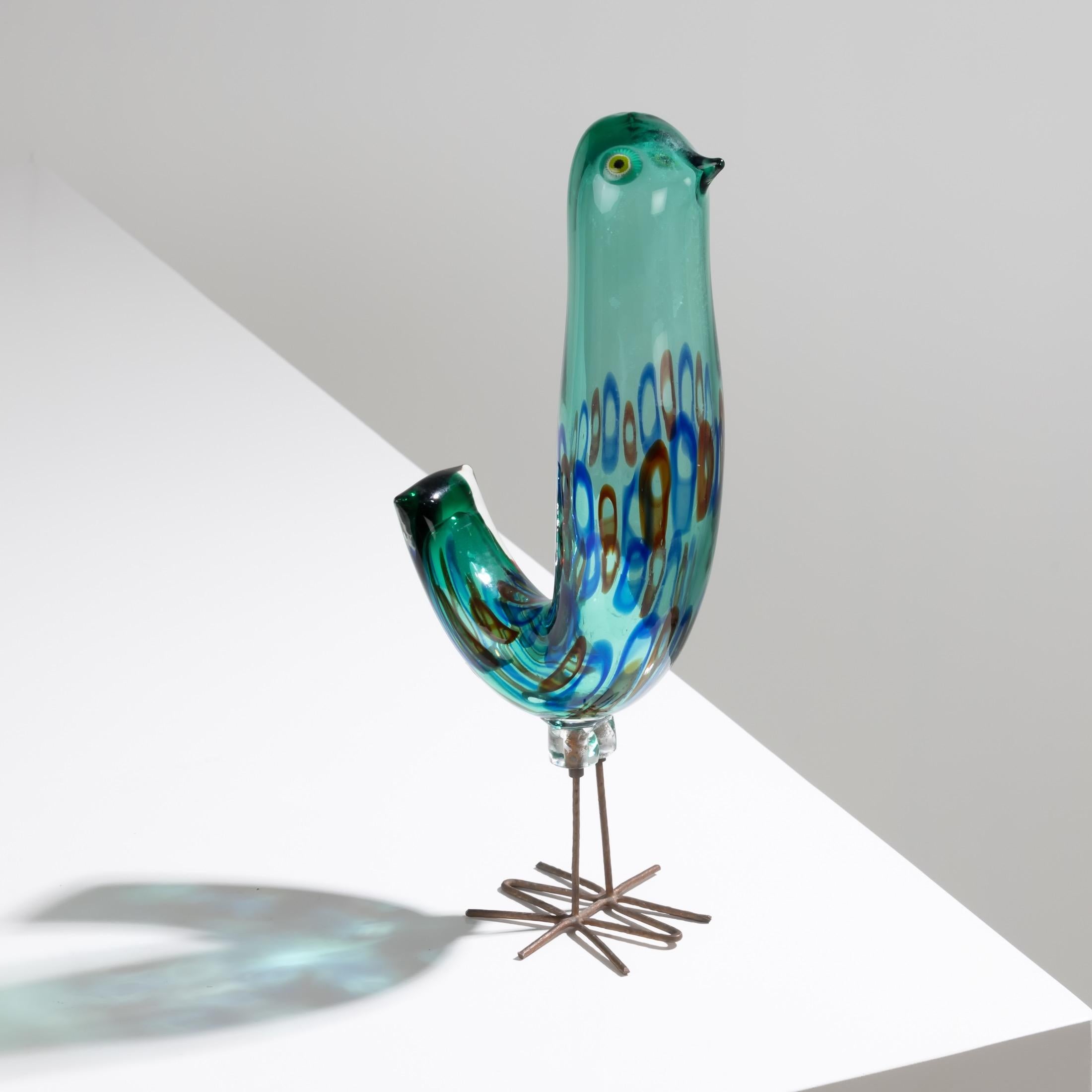 Glass bird resting on its hammered copper legs, designed by Alessandro Pianon for Vistosi in Murano, in 1962.
Its green glass body is decorated with red and blue murrines. its eyes are made of hot-applied murrines.
The object is listed in the