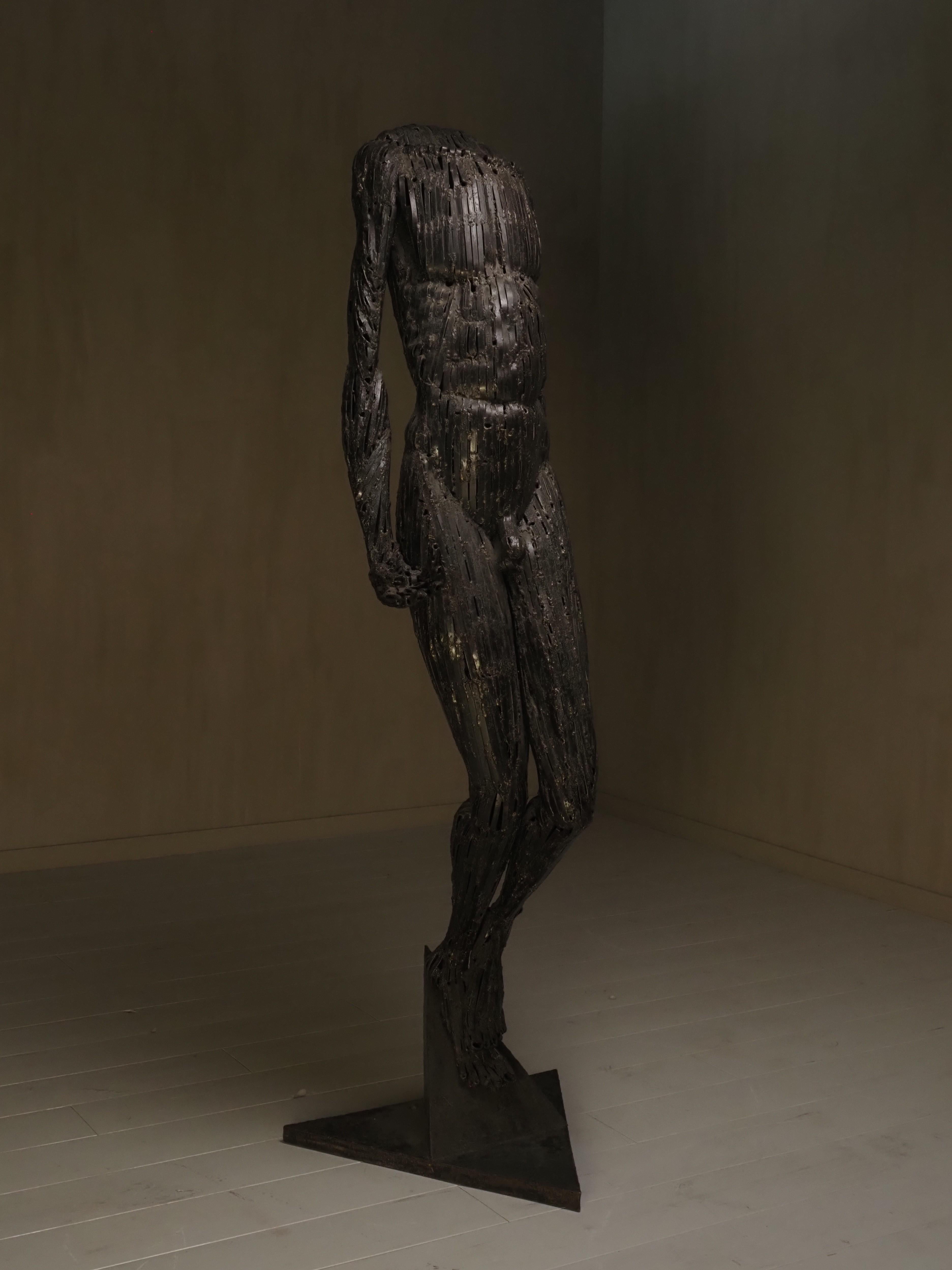 Questa sorprendente scultura contemporanea raffigura una figura maschile acefala in posizione contrapposta, catturata con un'elegante inclinazione in avanti. Realizzata interamente con frammenti di metallo scuro e saldato, la figura è grezza ma