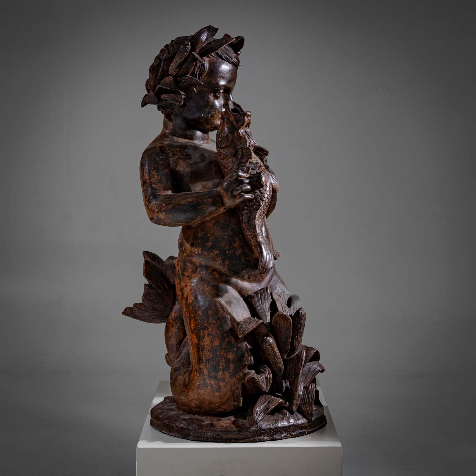 La sculpture d'un garçon agenouillé sur un piédestal rond devant une plante, tenant un poisson. Sa tête est ornée d'une couronne de laurier. La queue de poisson du garçon de mer est enroulée dans le dos. La sculpture est polyvalente. Diamètre de la