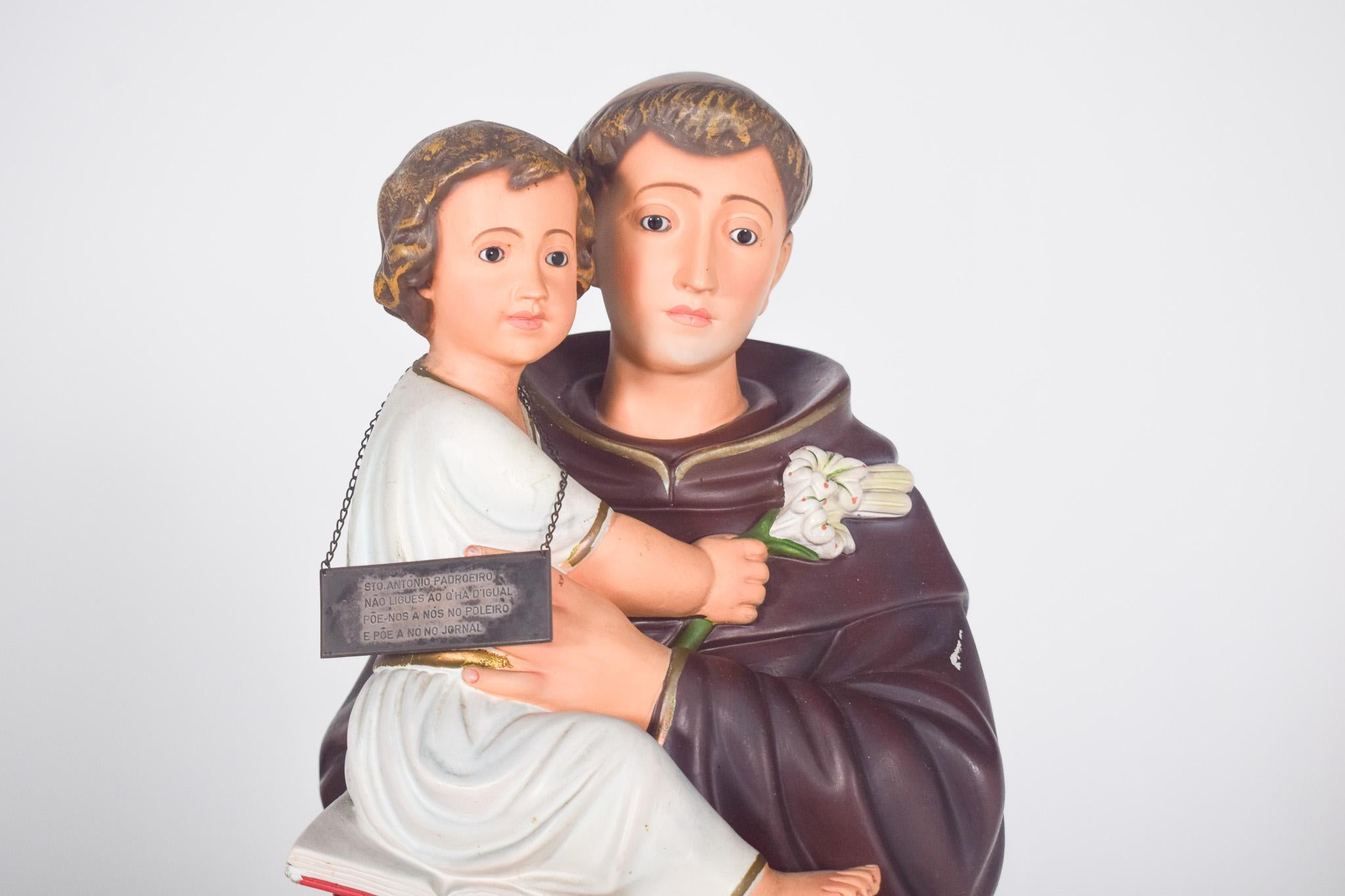 Sculpture of St Anthony with Child. St Anthony is depicted wearing a brown cloak, typical of the Franciscans, with gold details and a cord around his waist. The Child Jesus is dressed in white and gold and sitting on a book, holding a white lily in