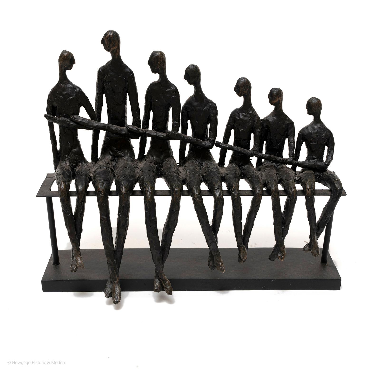 A group of 7 stick-thin figures sitting on a bench, each looking at the figure beside them and joining left hands with each other, in the manner of Alberto Giacometti.

Alberto Giacometti is best known for his elongated figures, often walking or