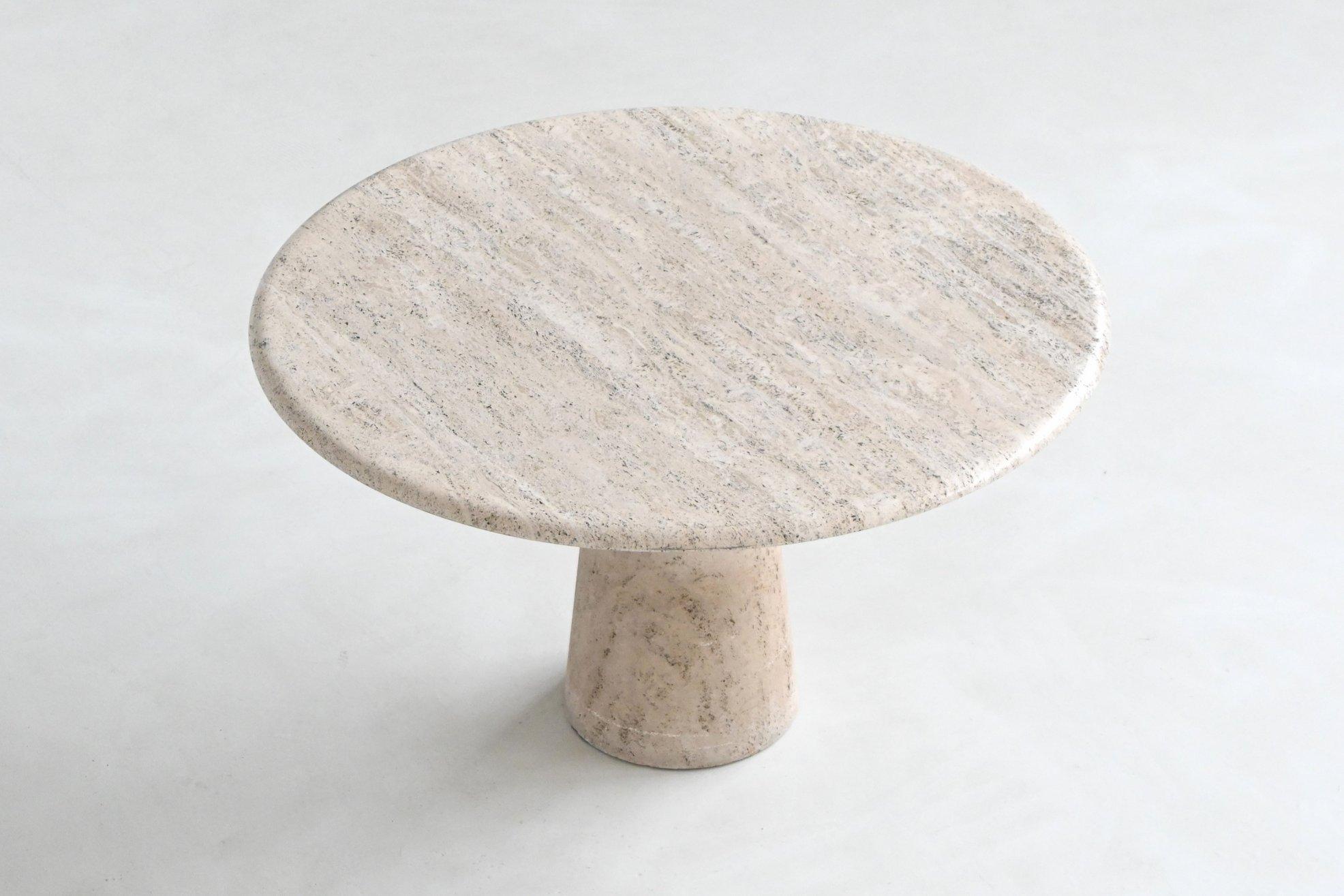 This sculptural dining table features a beautifully honed round travertine top with rich natural patterning in warm cream and soft beige tones. The surface displays a striking mix of veins, pores, and subtle color variations characteristic of