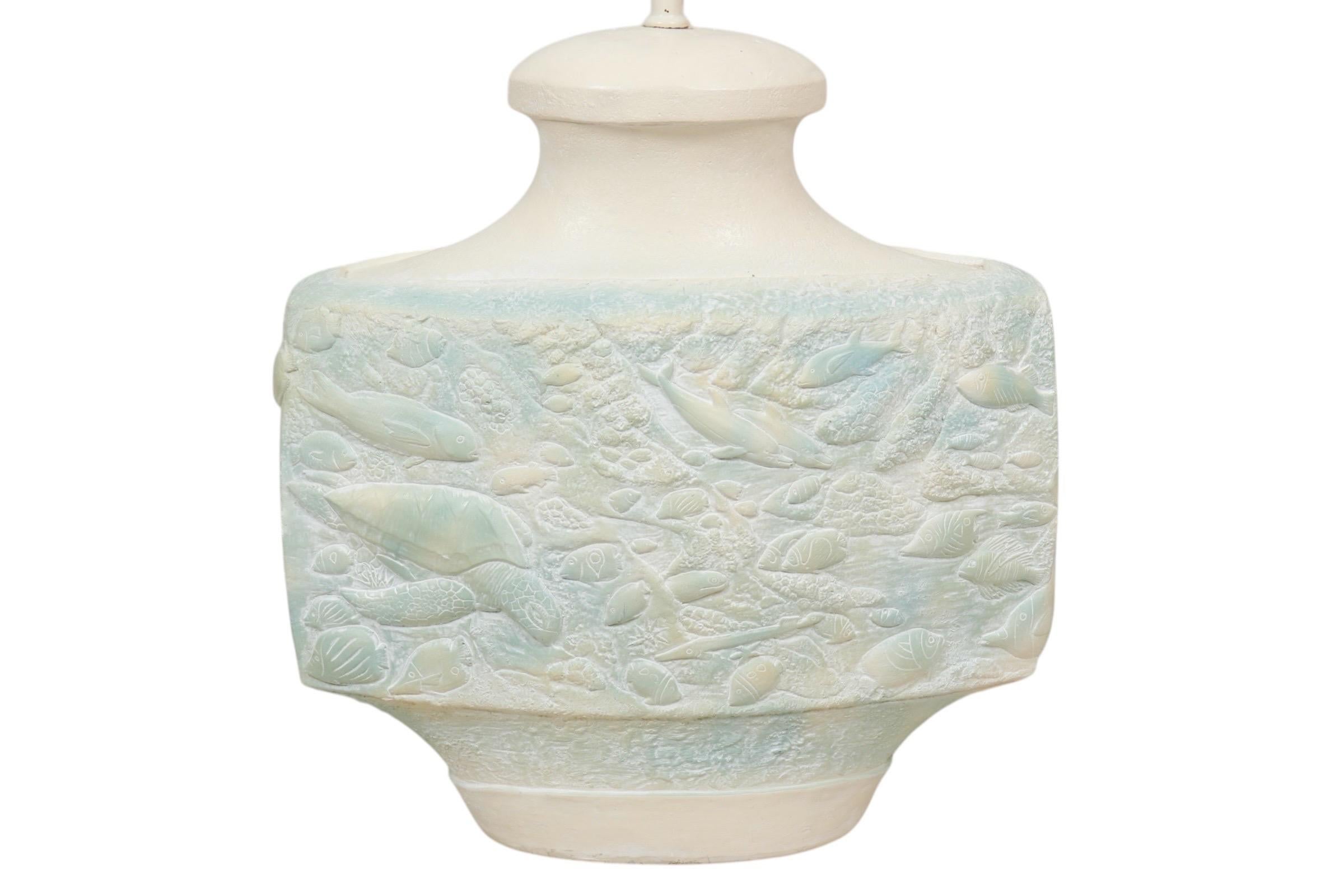 A coastal style cream and teal ceramic table lamp decorated with an under the waves sea life pattern in relief. The body is a rounded rectangular shape, embossed with loggerhead turtles, dolphins, lionfish, angel fish and others swimming amongst sea