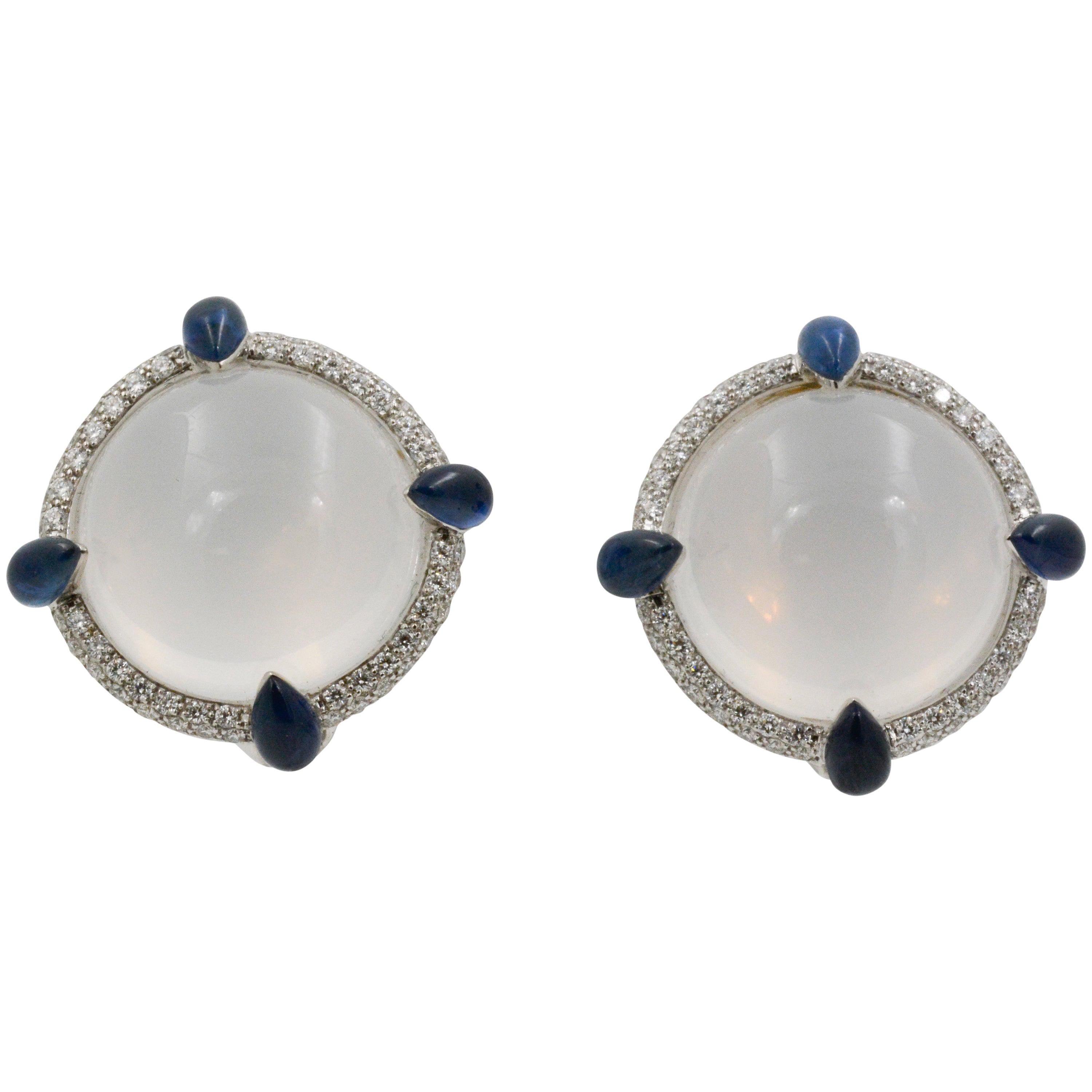 These Seaman Schepps 18k white gold earrings feature two cabochon white quartz that are accented with eight cabochon pear shaped sapphires and a pave diamond rim. The blue sapphires have a total weight of 3.68 carats and the diamonds have a total