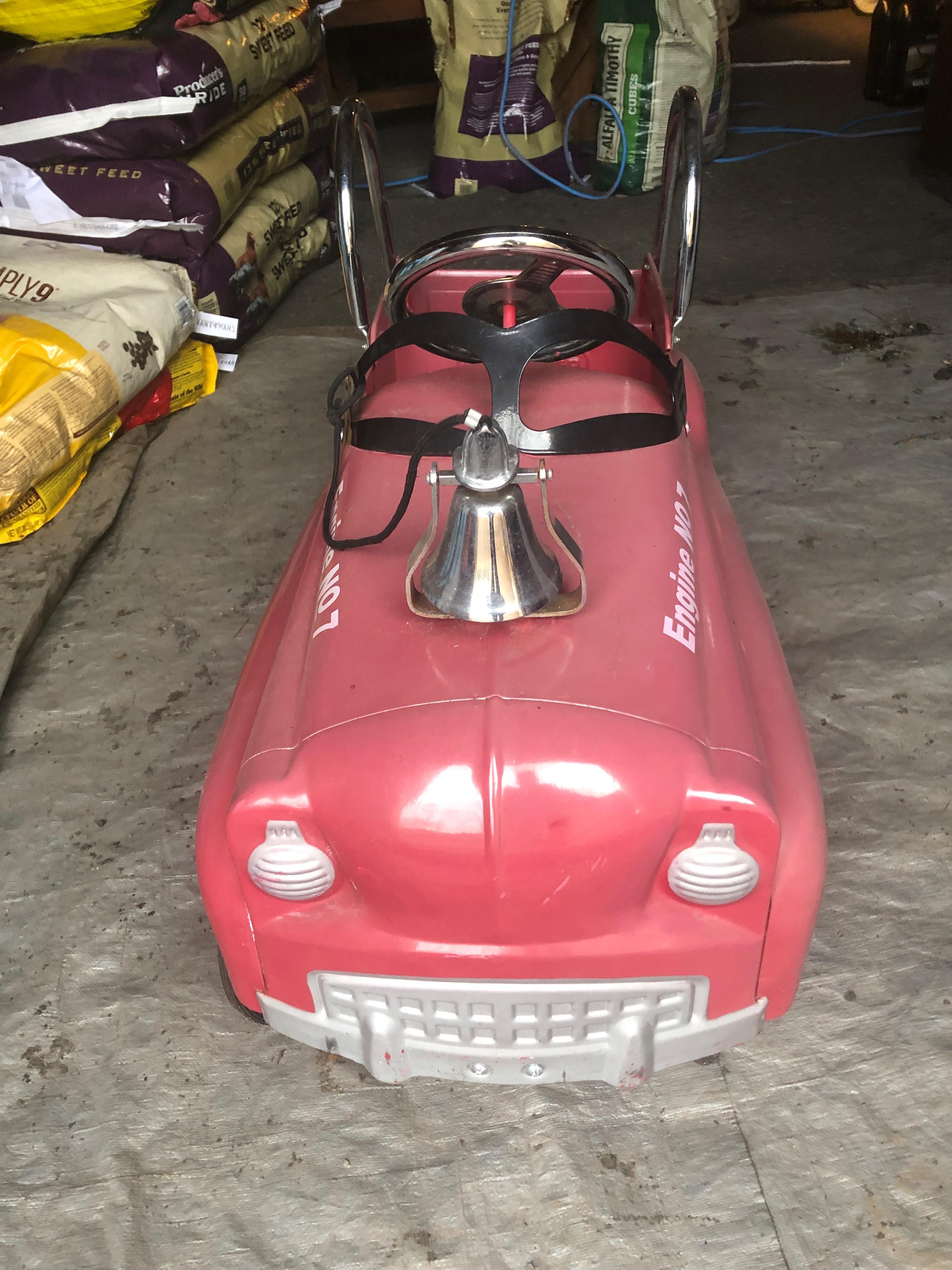 Ravissante voiture à pédales de pompier vintage dans une teinte claire de rouge et avec des lettres dorées de pompiers et de sauveteurs ainsi qu'un mignon chien dalmatien.