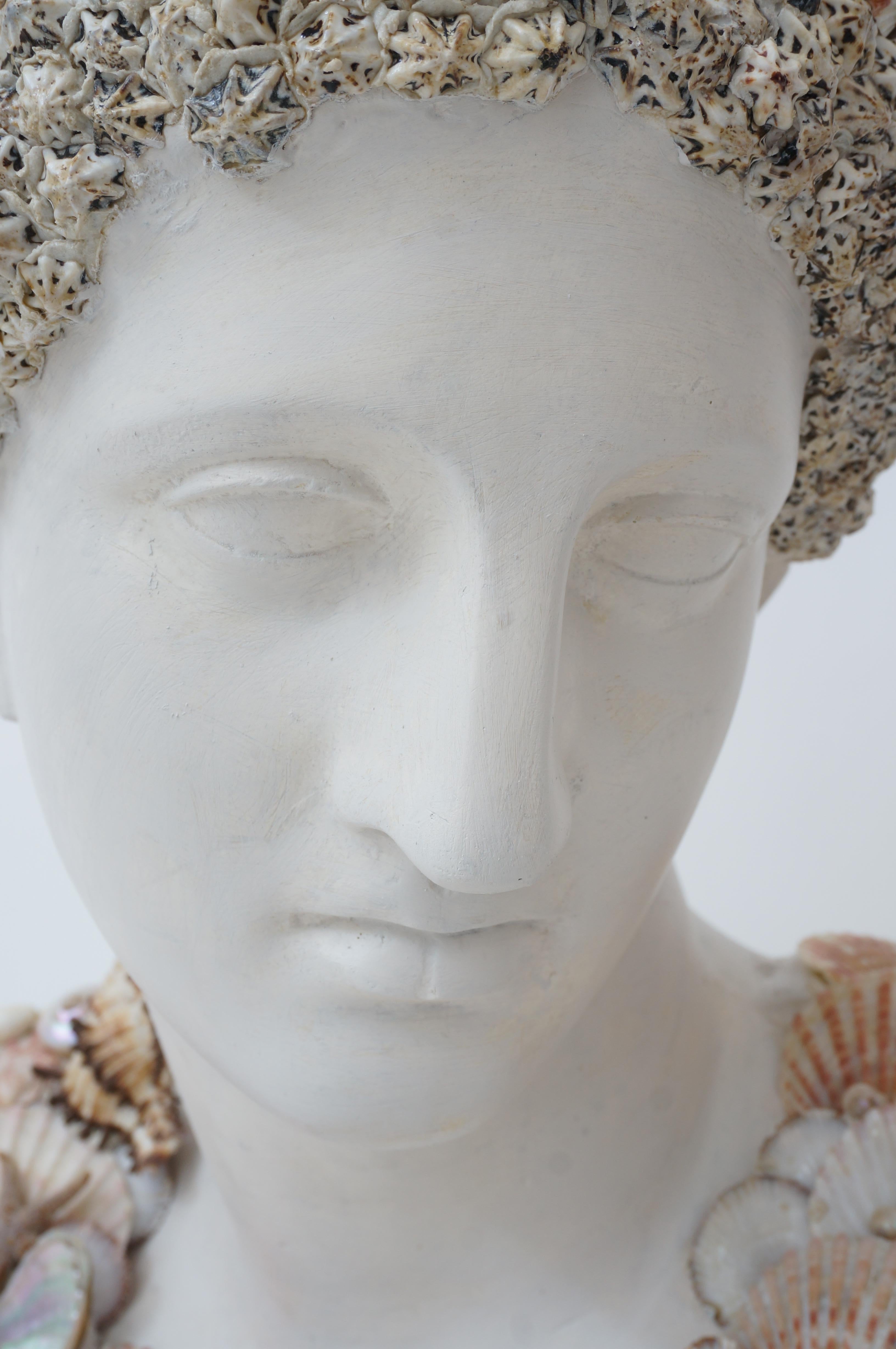 This chic neoclassical bust of a Greek goddess has been encrusted with seashells and is very much in the taste of pieces coveted by the iconic designer Tony Duquette. 

The piece has a painted faux marble finish on the base and the face a soft